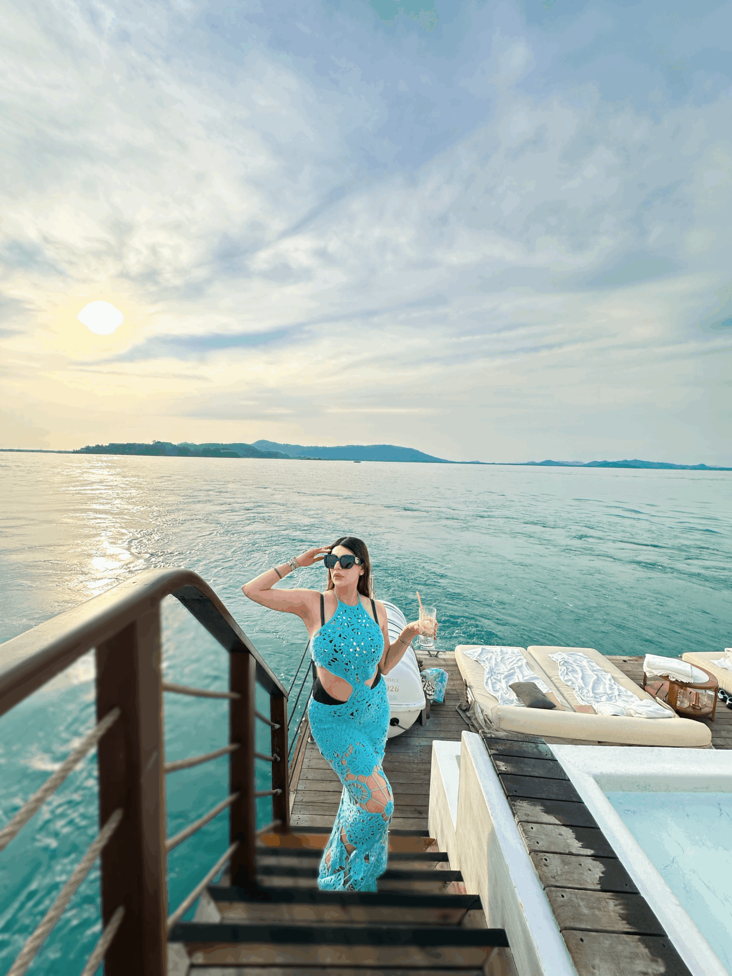 Woman in blue dress and sunglasses standing on stairs by water, with a boat and sky in background.