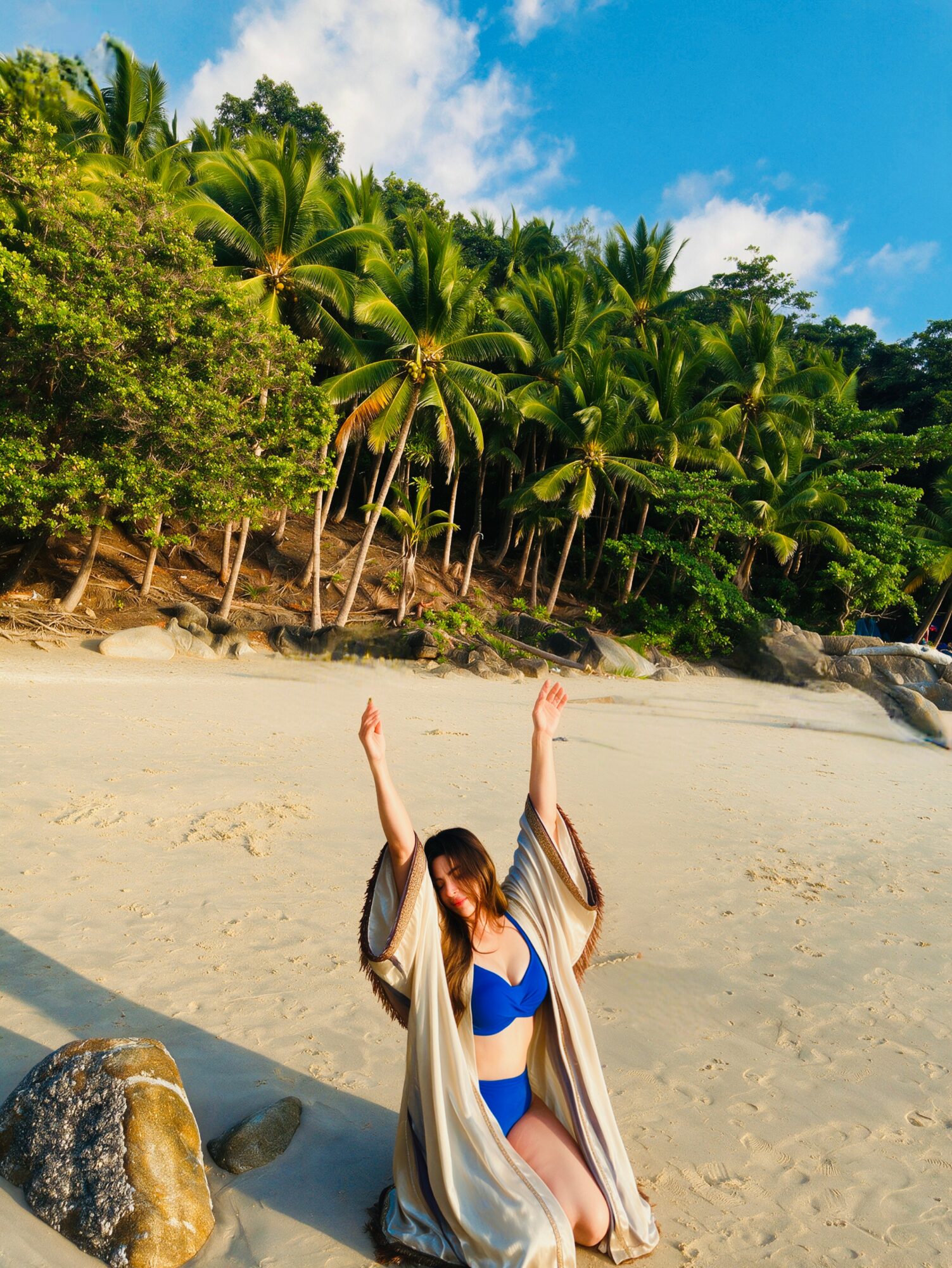 Person in blue bikini and beige cover-up sitting on sand with arms raised, tropical trees and blue sky in background.