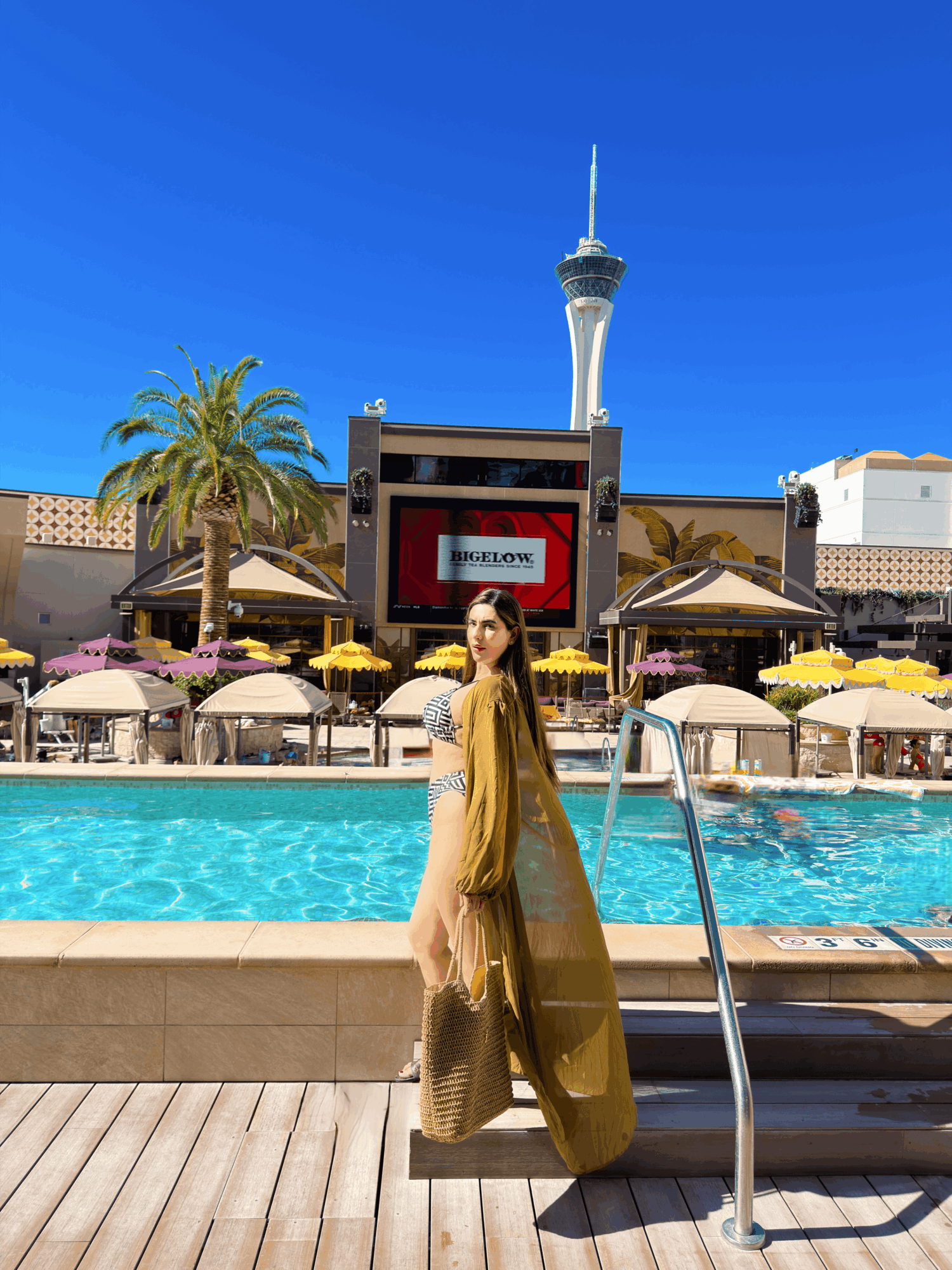 Woman in a yellow dress standing by a pool with a cityscape and tower in the background.