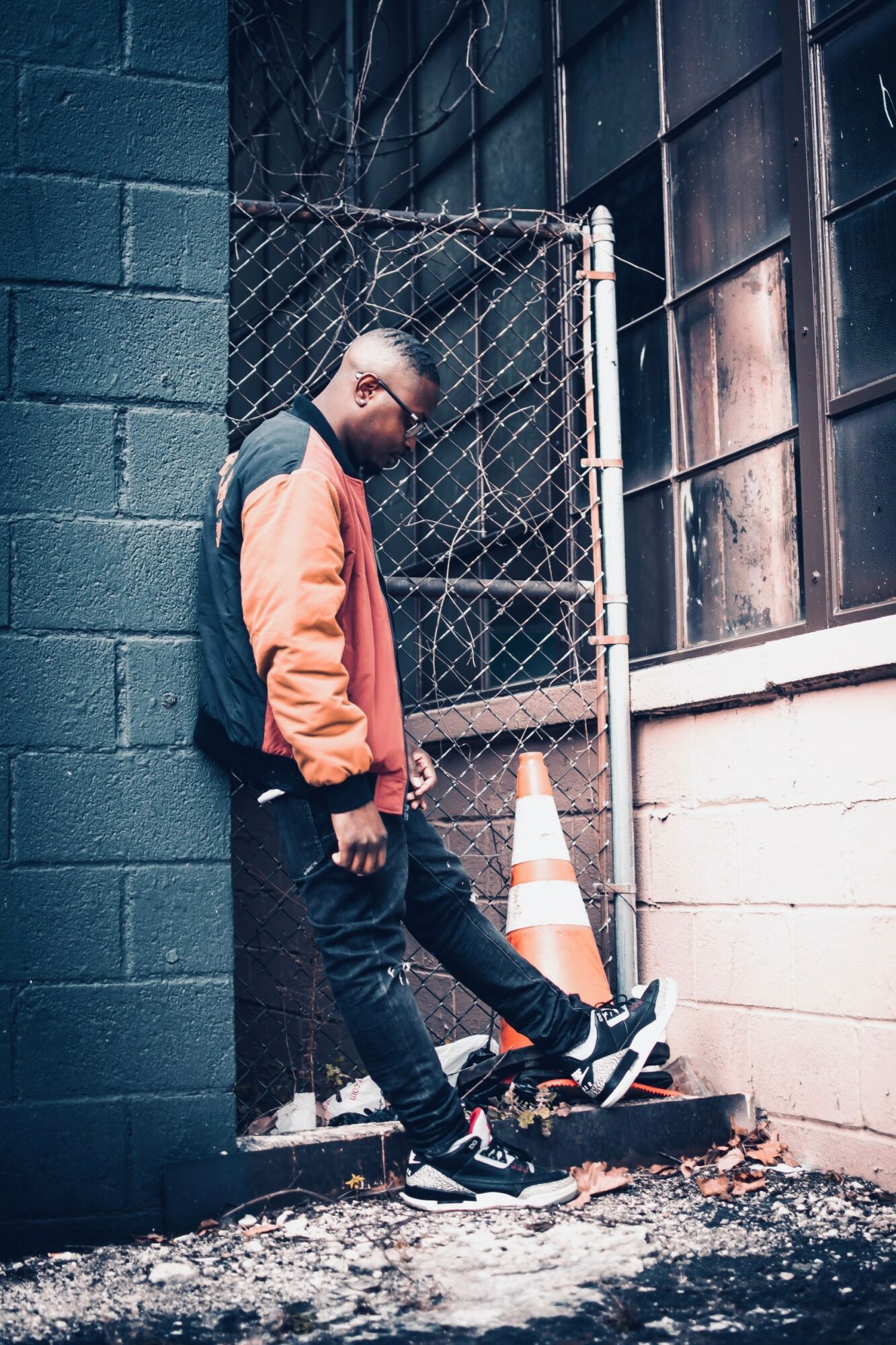 Person leaning against a chain-link fence in an urban alleyway, wearing a jacket and sneakers, with a traffic cone nearby.