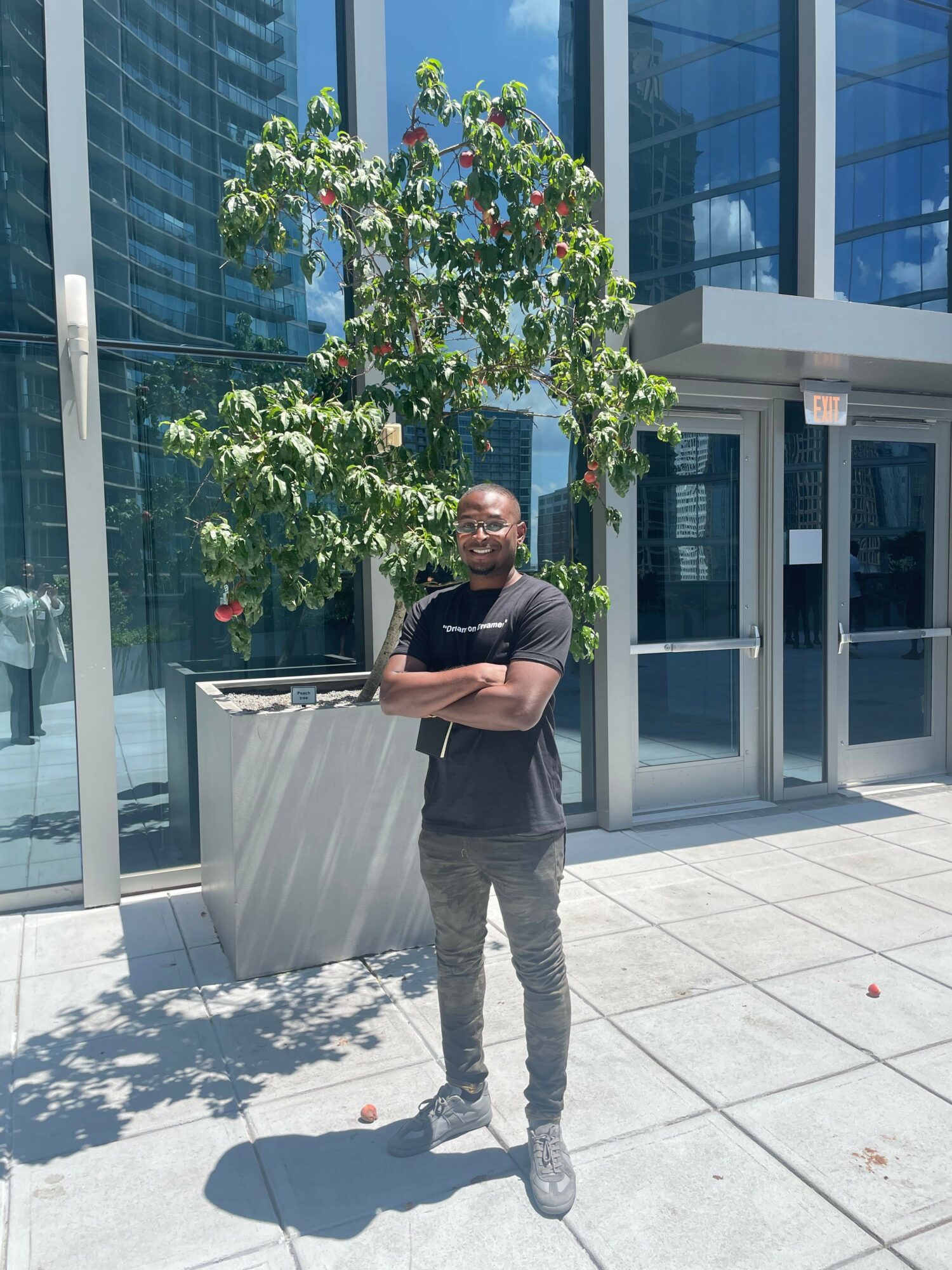 Man standing outside near a tree and glass building, holding a tablet, with shadows on the ground.