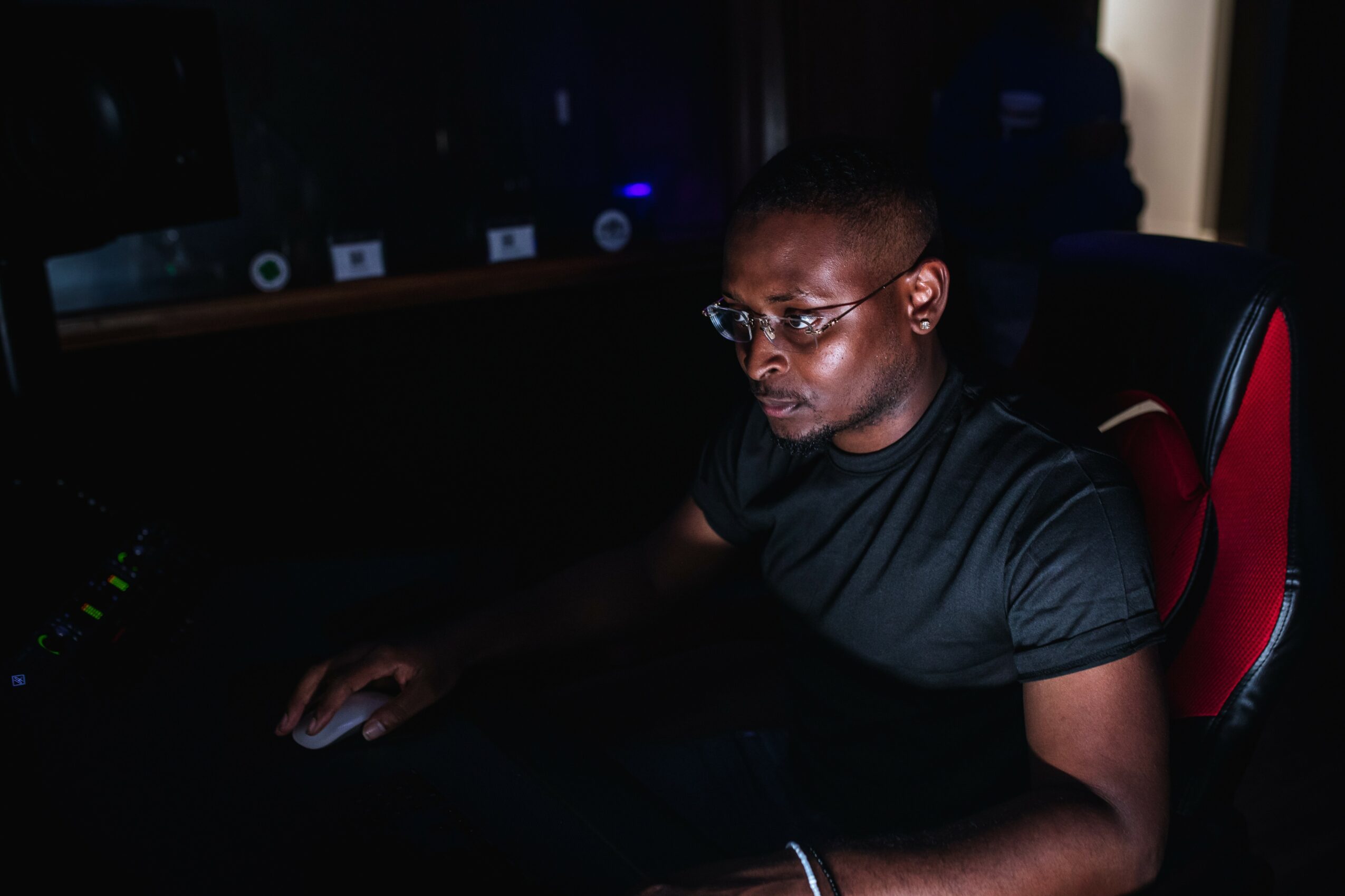 Person sitting in a dark room, using a computer mouse, wearing glasses and a black shirt.