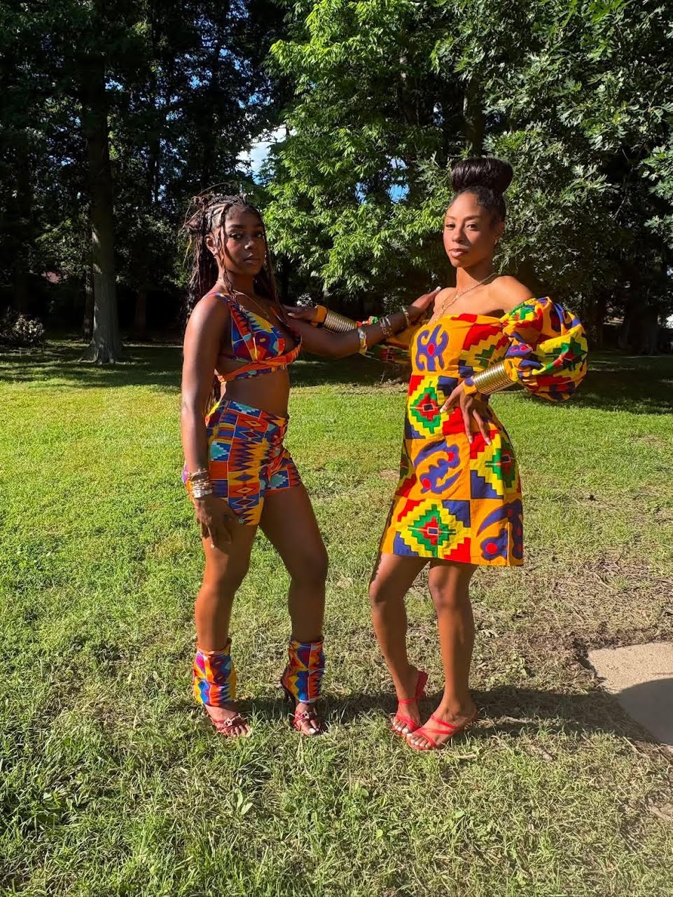 Two women in colorful African print outfits standing on grass in a park with trees in the background.