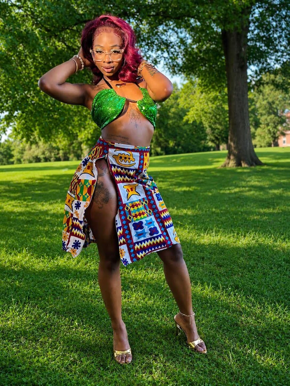 Woman with pink hair in a green top and colorful skirt outdoors on grass, trees in background.