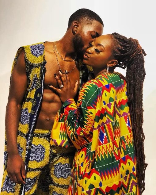 A man and woman are close, touching foreheads and eyes closed, wearing colorful clothing with patterns, in an intimate moment.