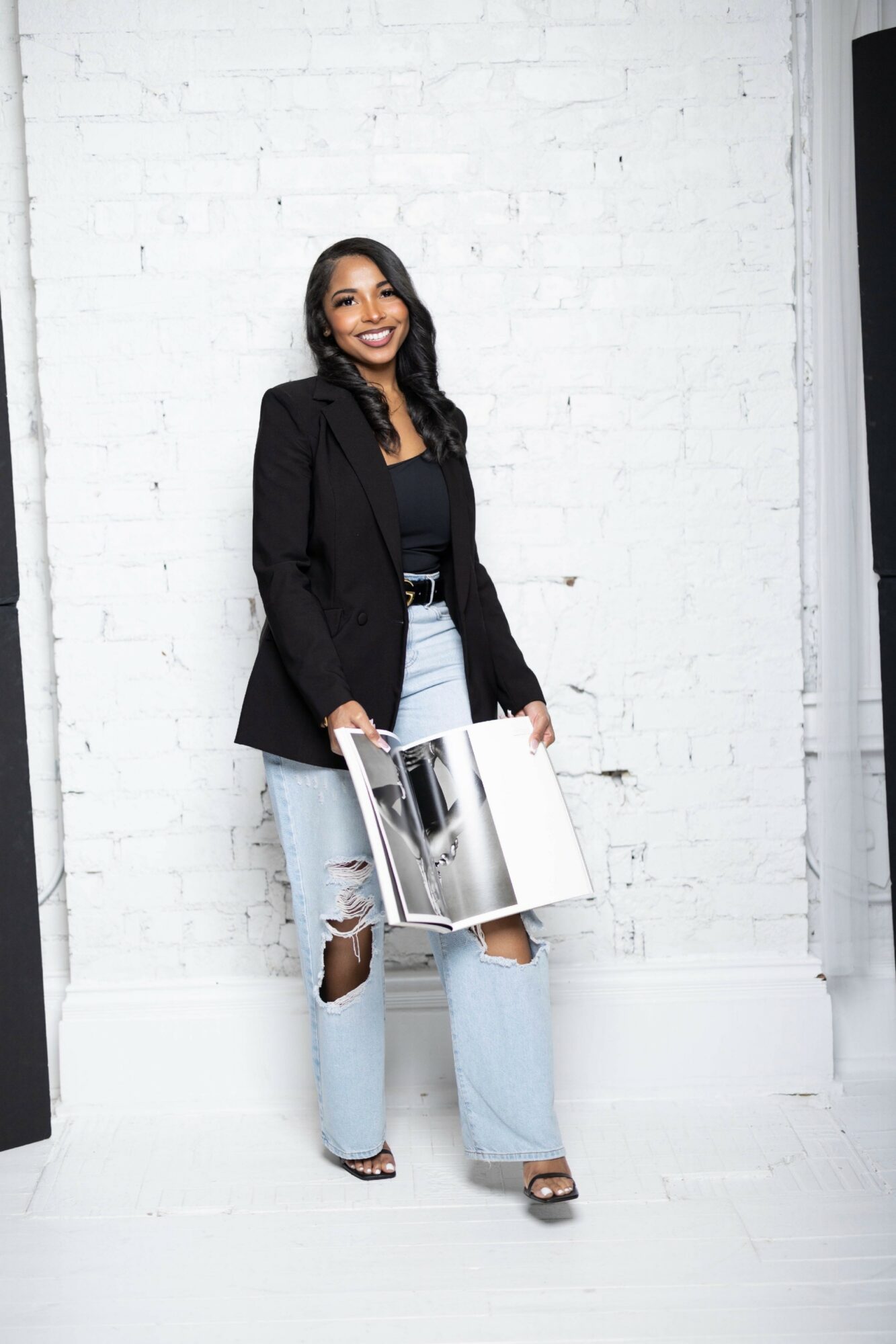 Woman with long dark hair smiling, holding an open magazine, standing against a white brick wall, wearing a black blazer and ripped jeans.