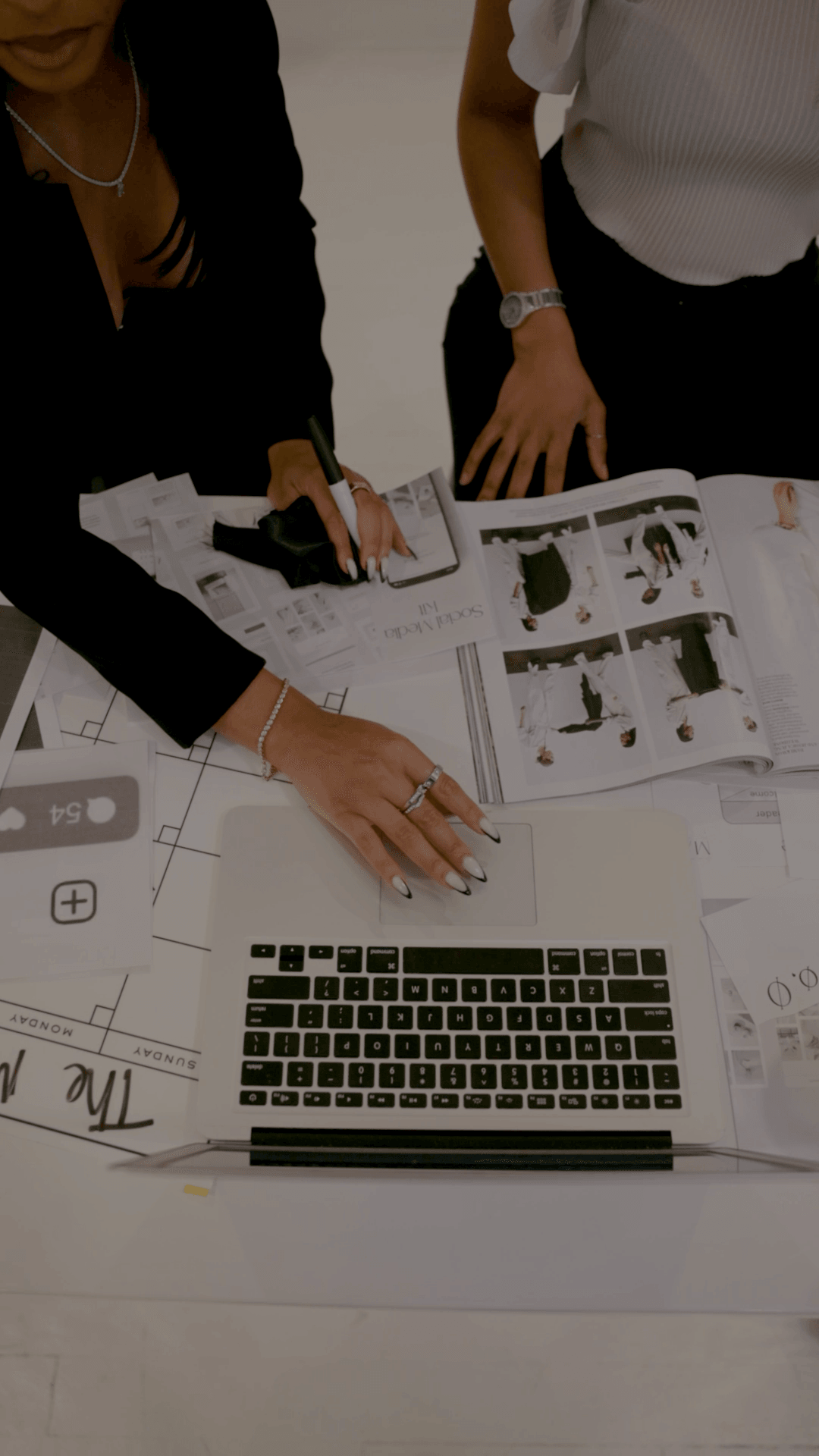 Two people work on documents and a laptop on a table, with papers and charts spread out.