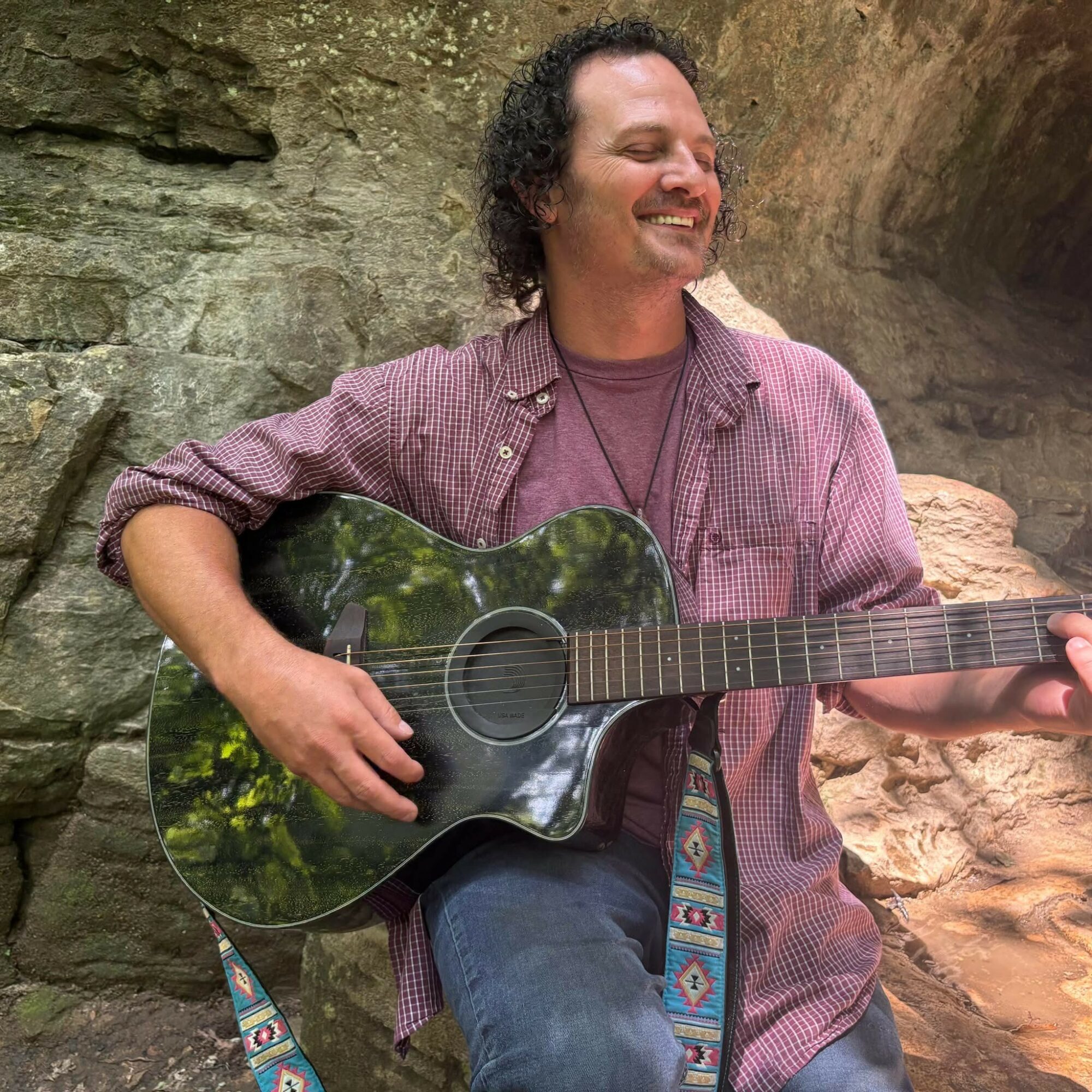Person smiling and playing guitar outdoors near rocks, wearing a checkered shirt and jeans.