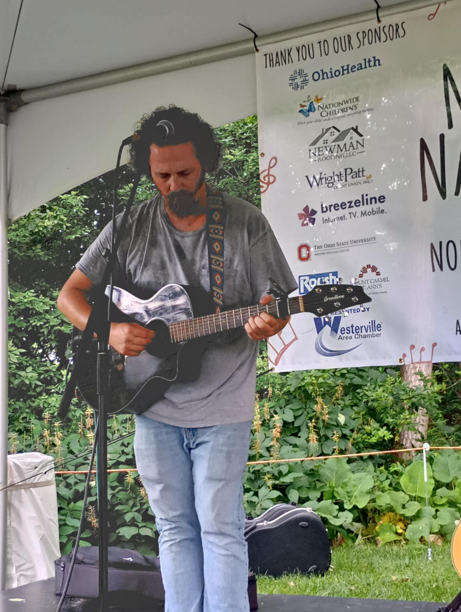 Man with curly hair and beard playing guitar on stage outdoors, with a banner and greenery in background.