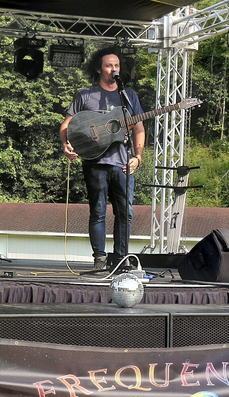 Man with a guitar performing on an outdoor stage with trees in the background.