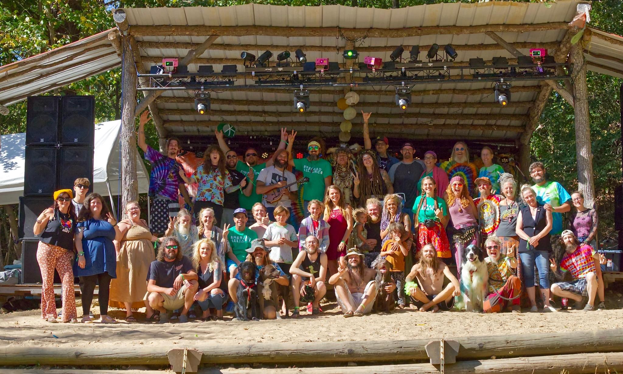 Group of people on stage outdoors, some dancing and posing, with trees and stage equipment visible behind them.