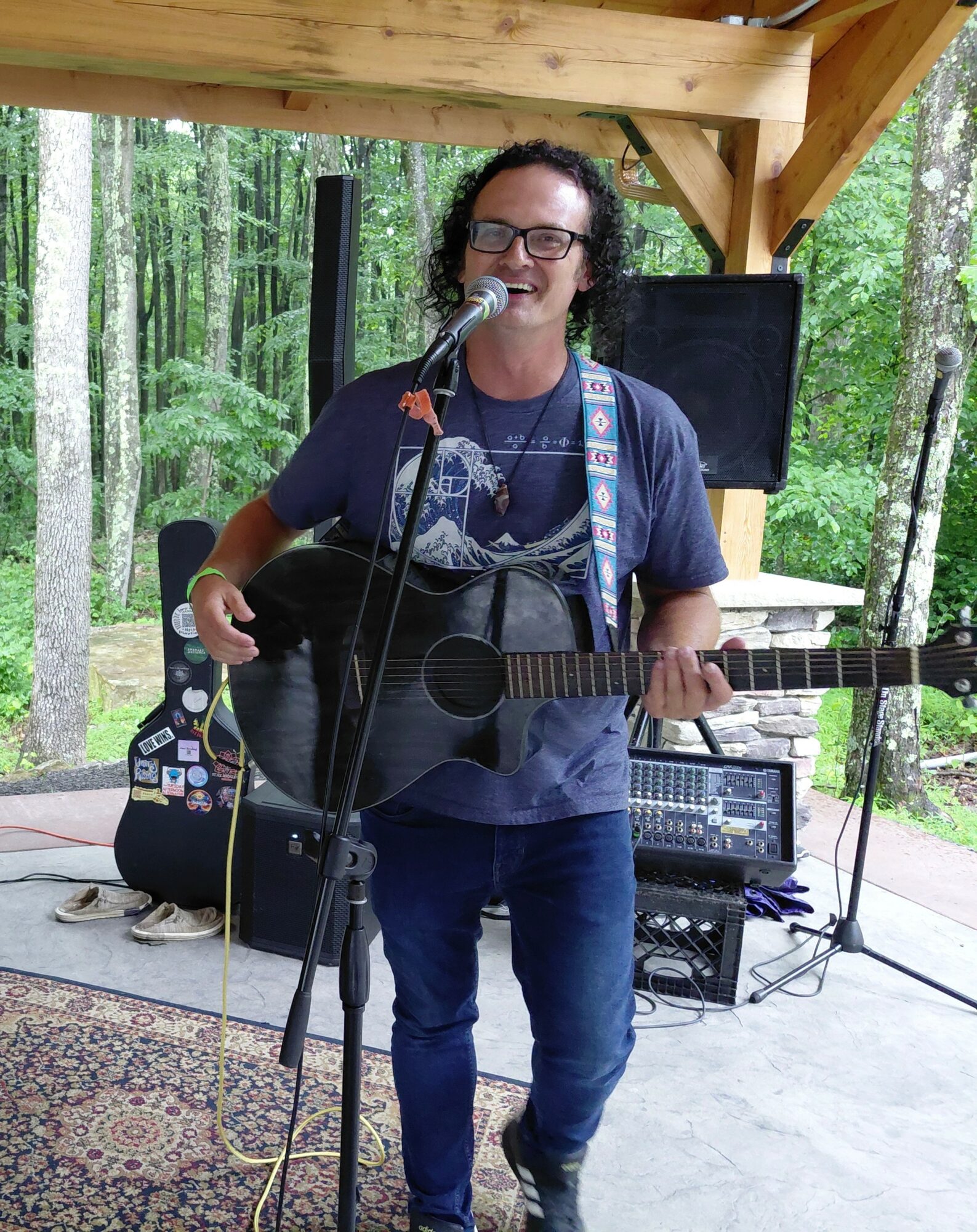Person with curly hair and glasses singing into a microphone while playing an acoustic guitar outdoors under a wooden shelter.