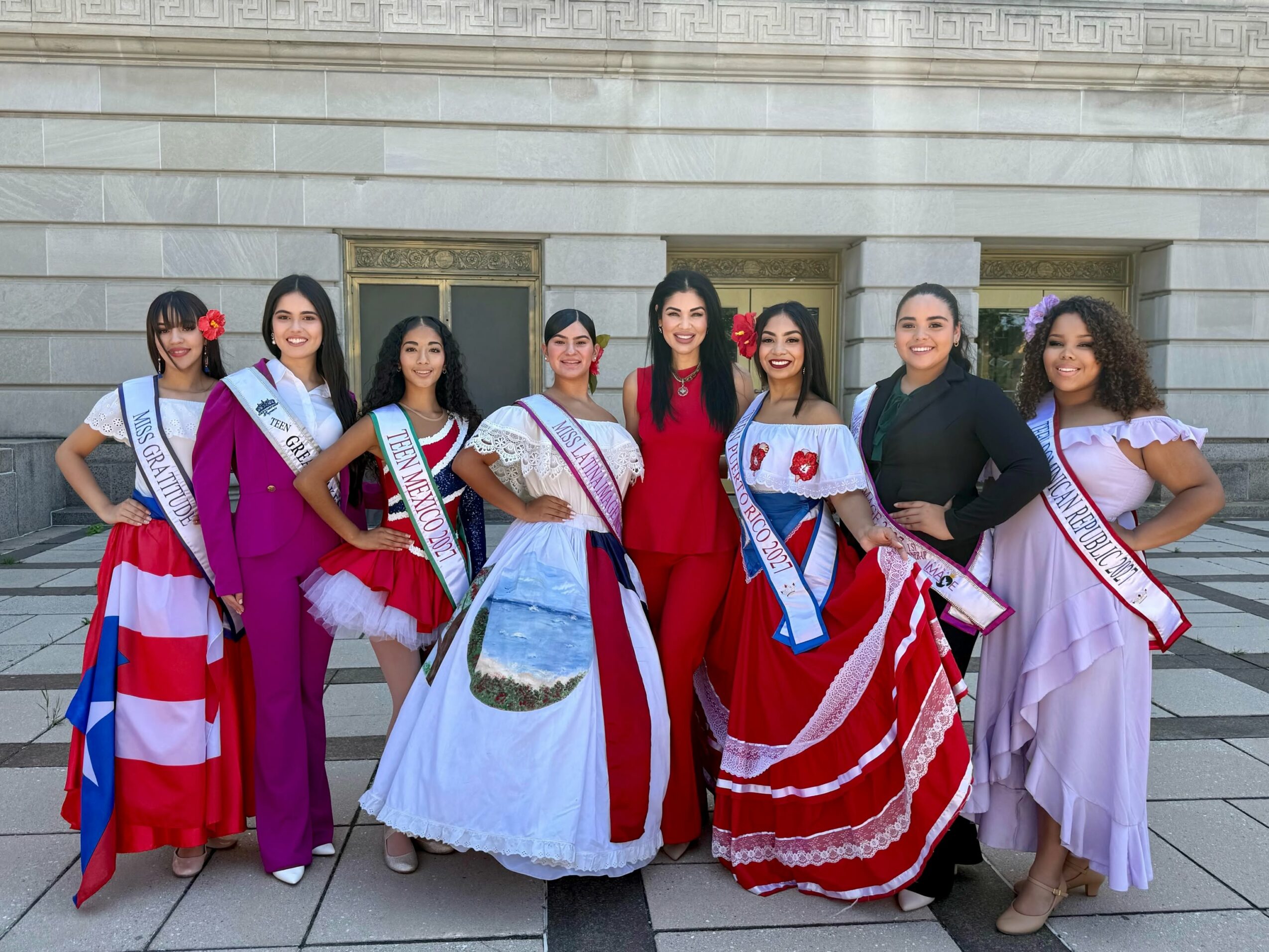 Group of women in colorful dresses and sashes standing outside a building with columns.