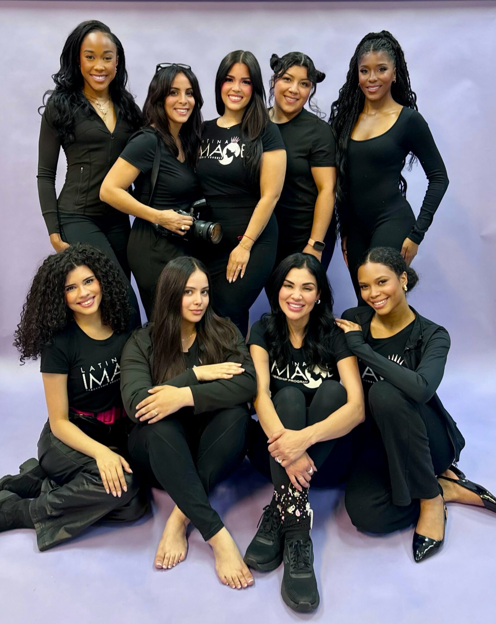 Group of nine women posing together, some standing and some sitting, smiling, in black clothing against a light background.