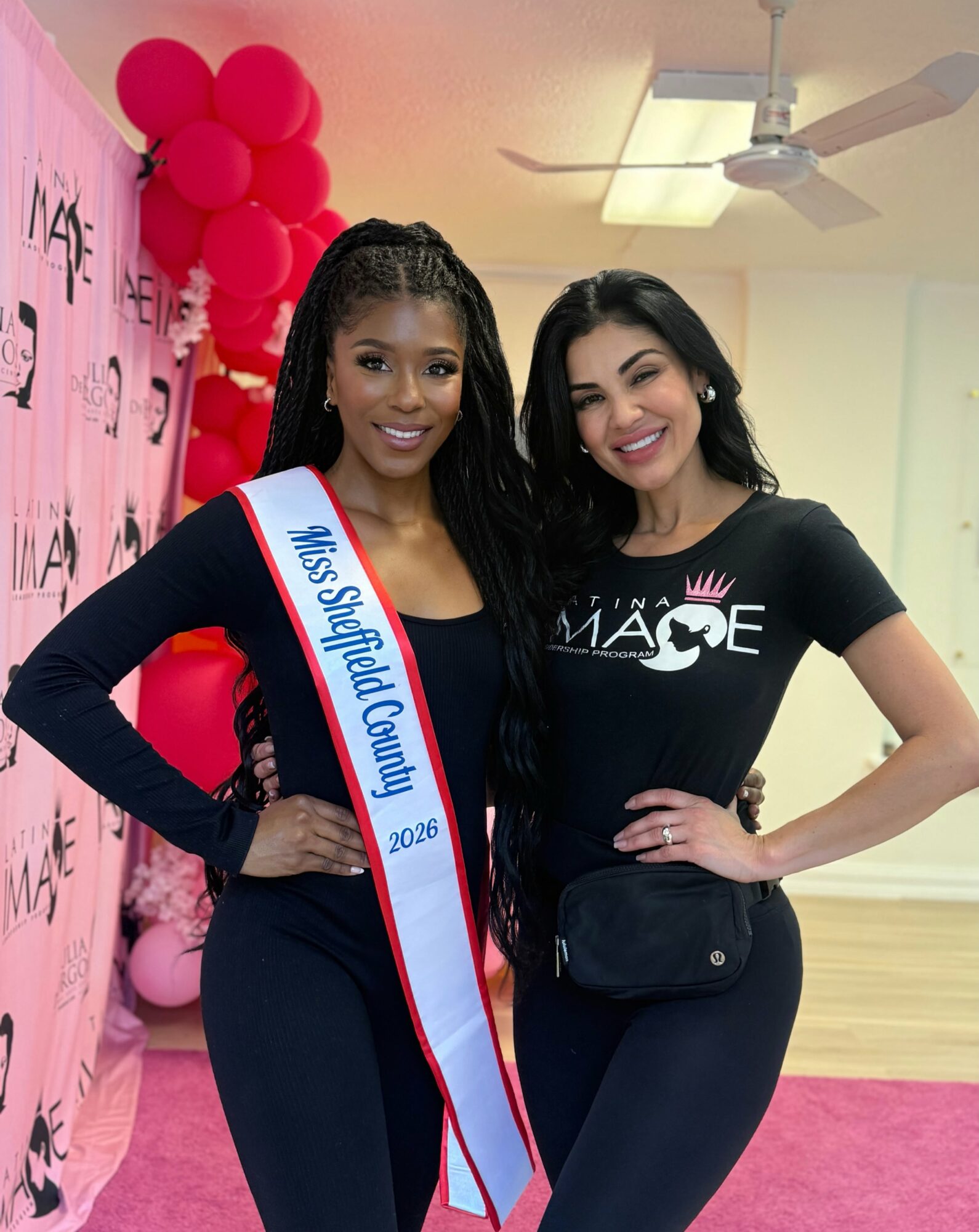Two women smiling, one wearing a sash that reads 'Miss Stratford County 2023,' in a decorated room with pink balloons and a pink carpet.