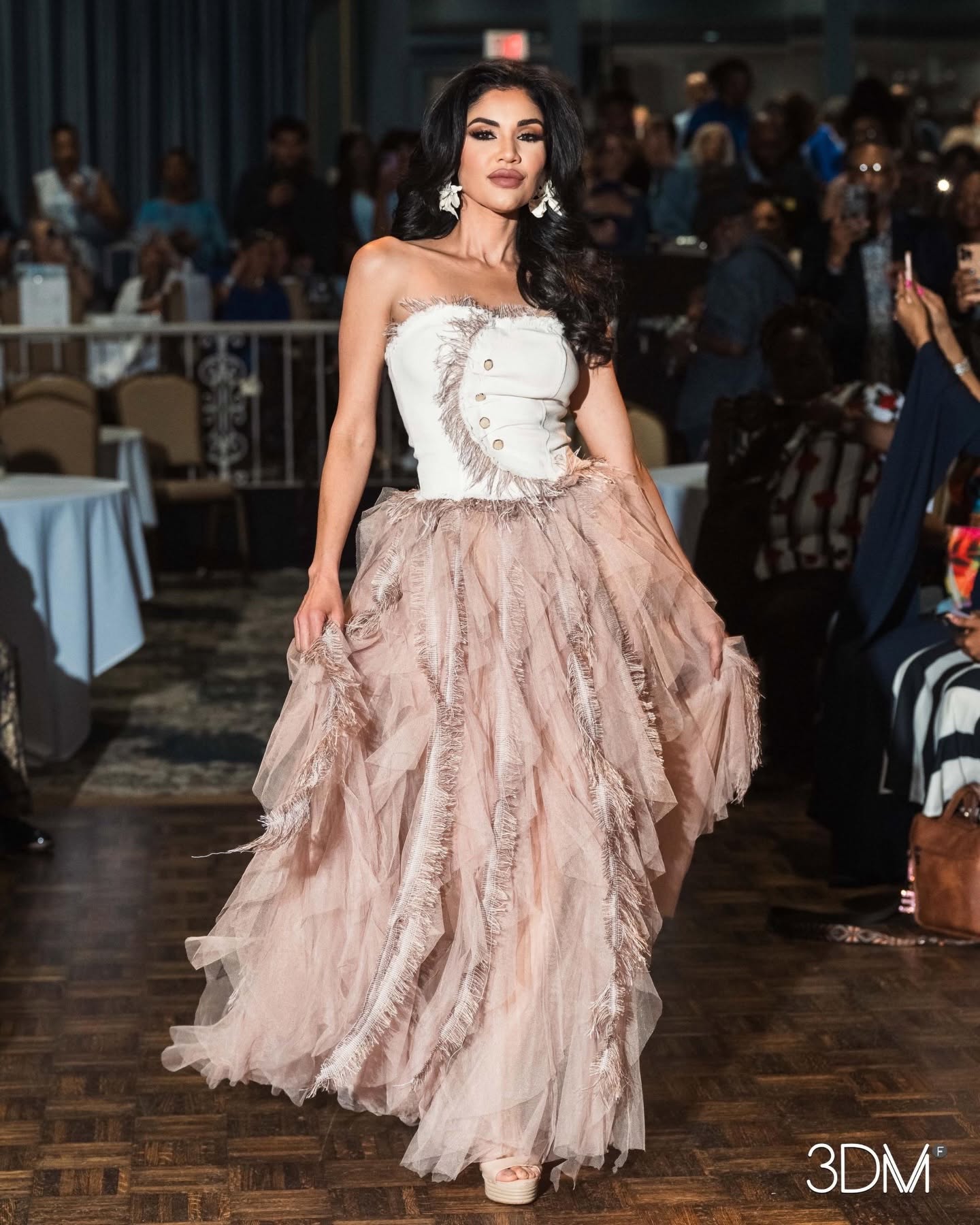 Woman in strapless white top and layered pink tulle skirt walking on a wooden floor at an event, with people in the background.