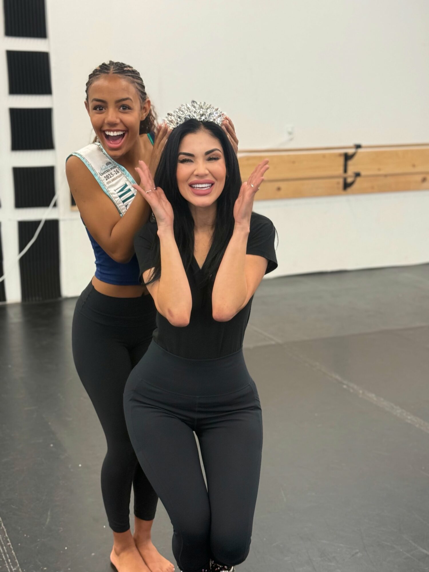 Two women smiling, one wearing a tiara, in a dance studio with a wooden barre, celebrating together.