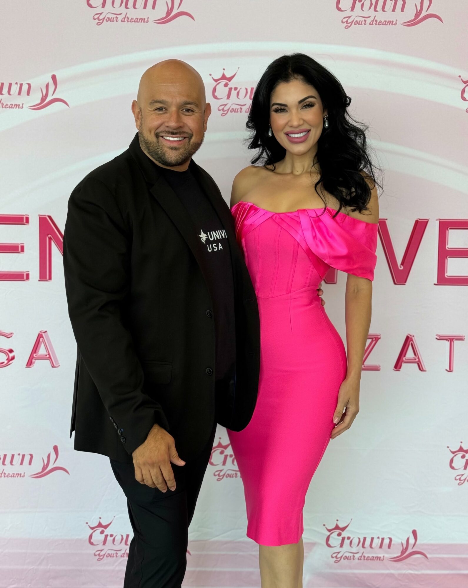 Man and woman standing together, smiling, in front of a pink and white backdrop with text and logos.