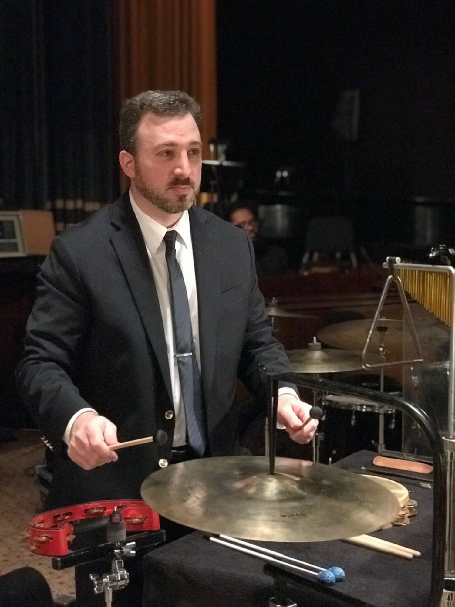 Man in suit playing percussion instrument with drumsticks in a performance setting.