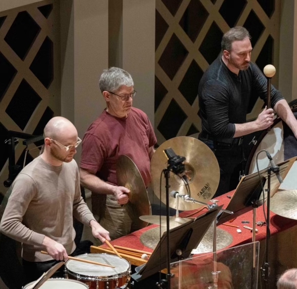 Three musicians playing percussion and a mallet instrument in a recording studio, with music stands and microphones.