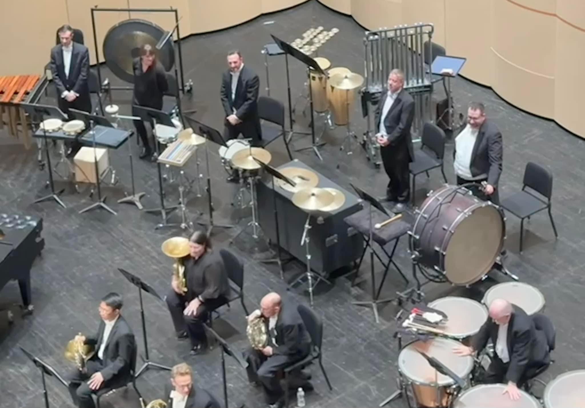 Orchestra members in formal attire with percussion instruments on stage, some seated and some standing, in a concert hall.