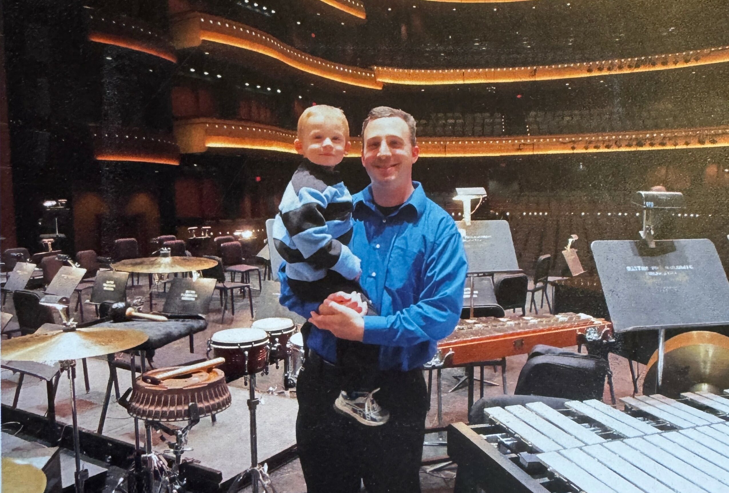 Man holding a child in a concert hall with musical instruments and stage equipment visible.