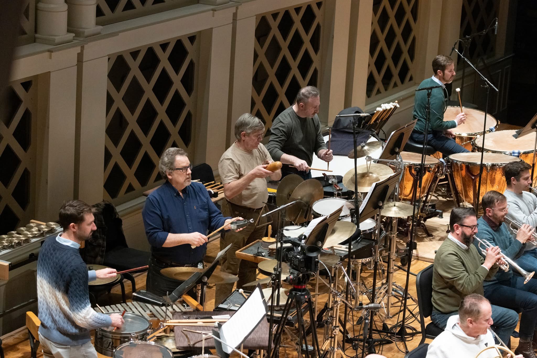 Orchestra performing on stage with musicians playing various instruments, including drums and wind instruments, in a concert hall.