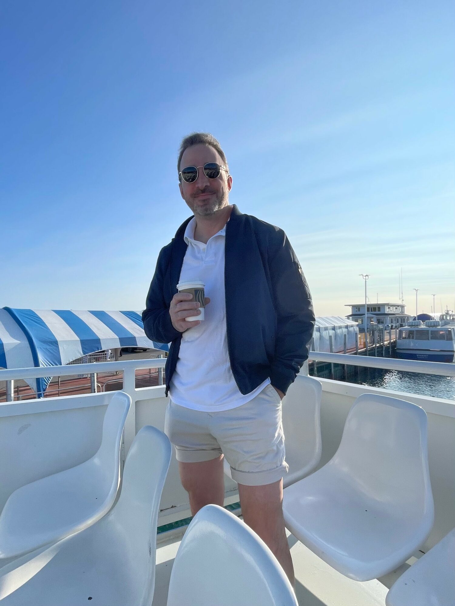 Man standing outdoors holding a drink, wearing sunglasses, a white shirt, navy jacket, and light-colored shorts, with a marina in background.