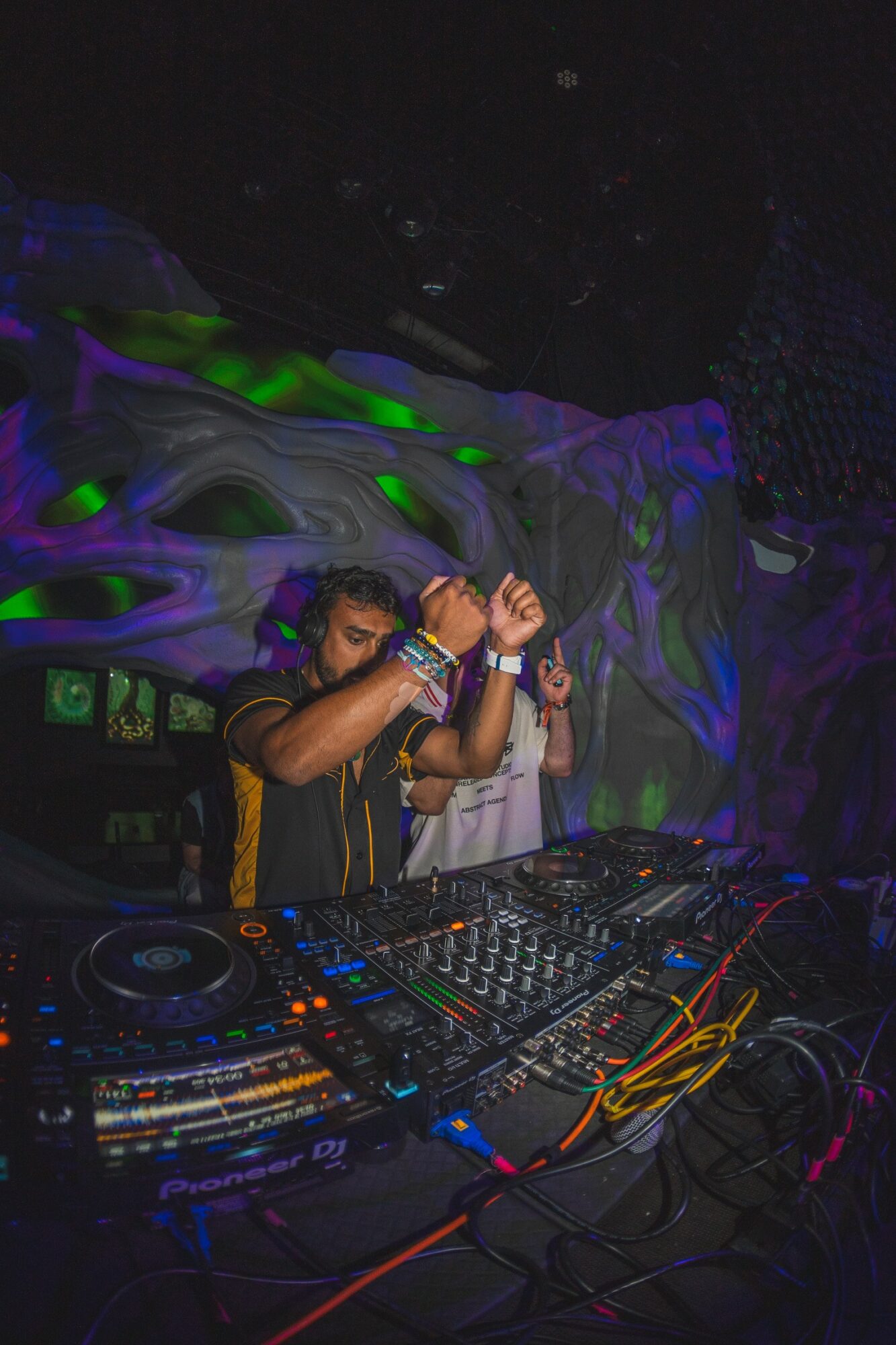 DJ wearing headphones and a black shirt with yellow accents, mixing music at a club with colorful lighting and abstract background.