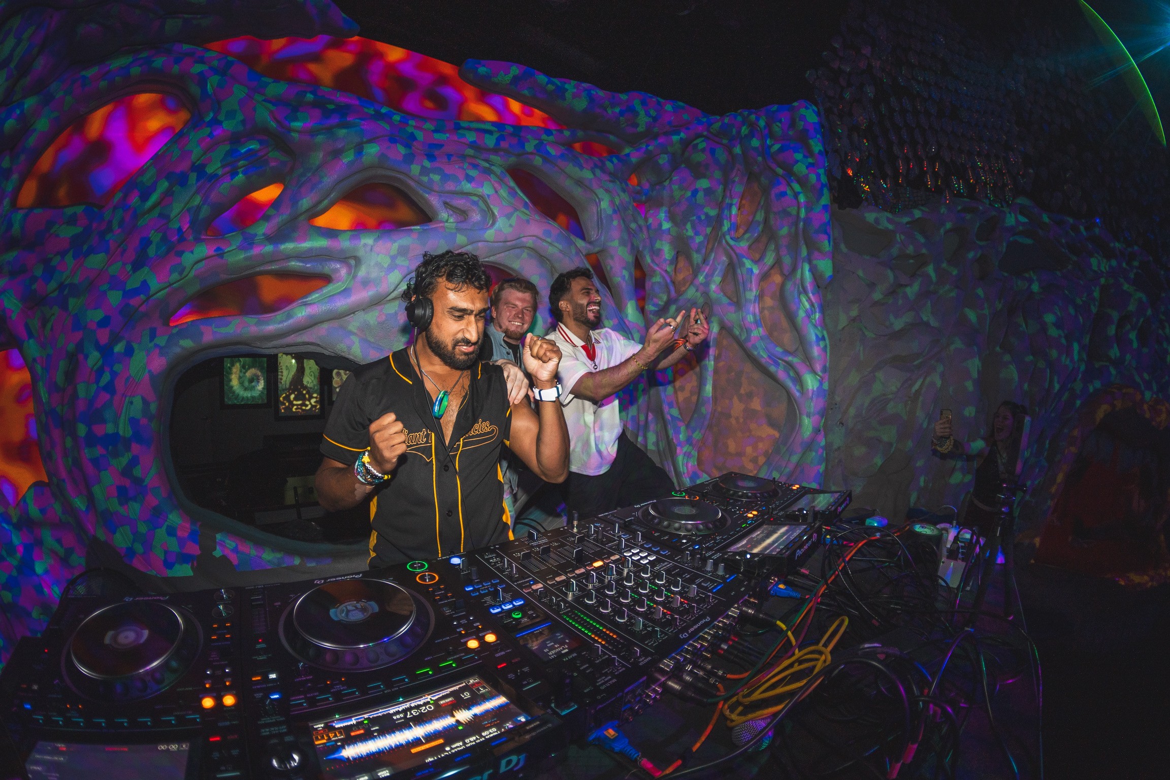 Two DJs perform in front of a colorful, psychedelic backdrop with electronic music equipment, in a dark club setting.