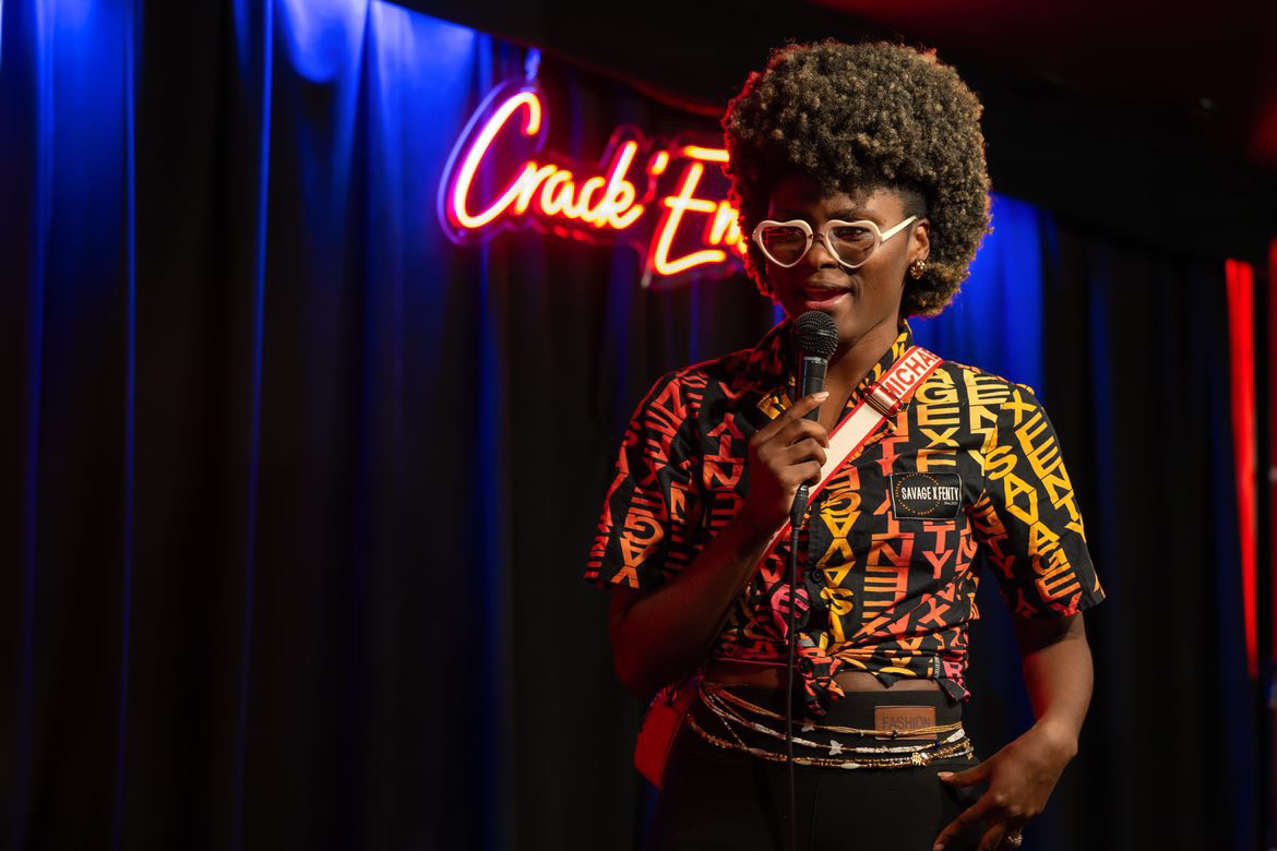 Person with curly hair and glasses speaking into a microphone on stage with a neon sign background