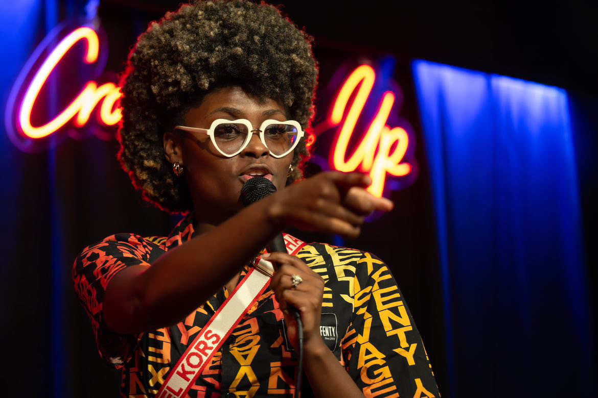 Woman with curly hair, glasses, and colorful clothing performs on stage with a microphone, neon sign in background.