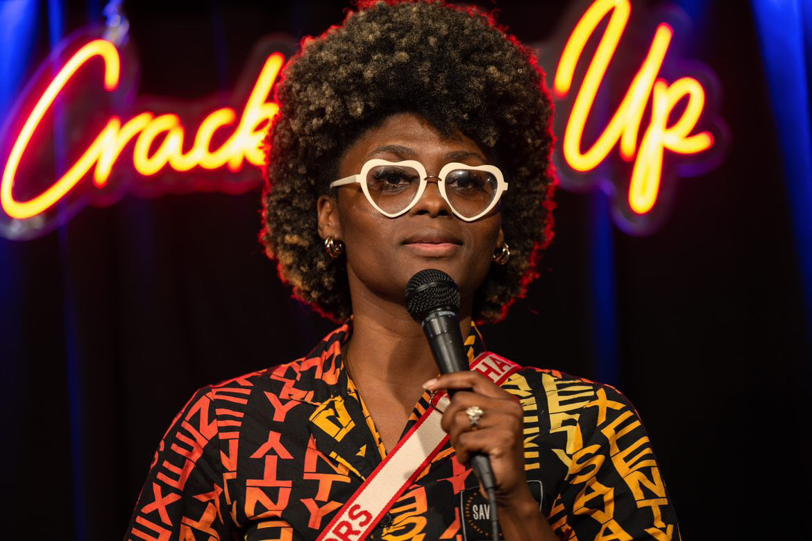 Woman with curly hair, glasses, holding a microphone, standing in front of a neon sign that says 'Crack Up'.