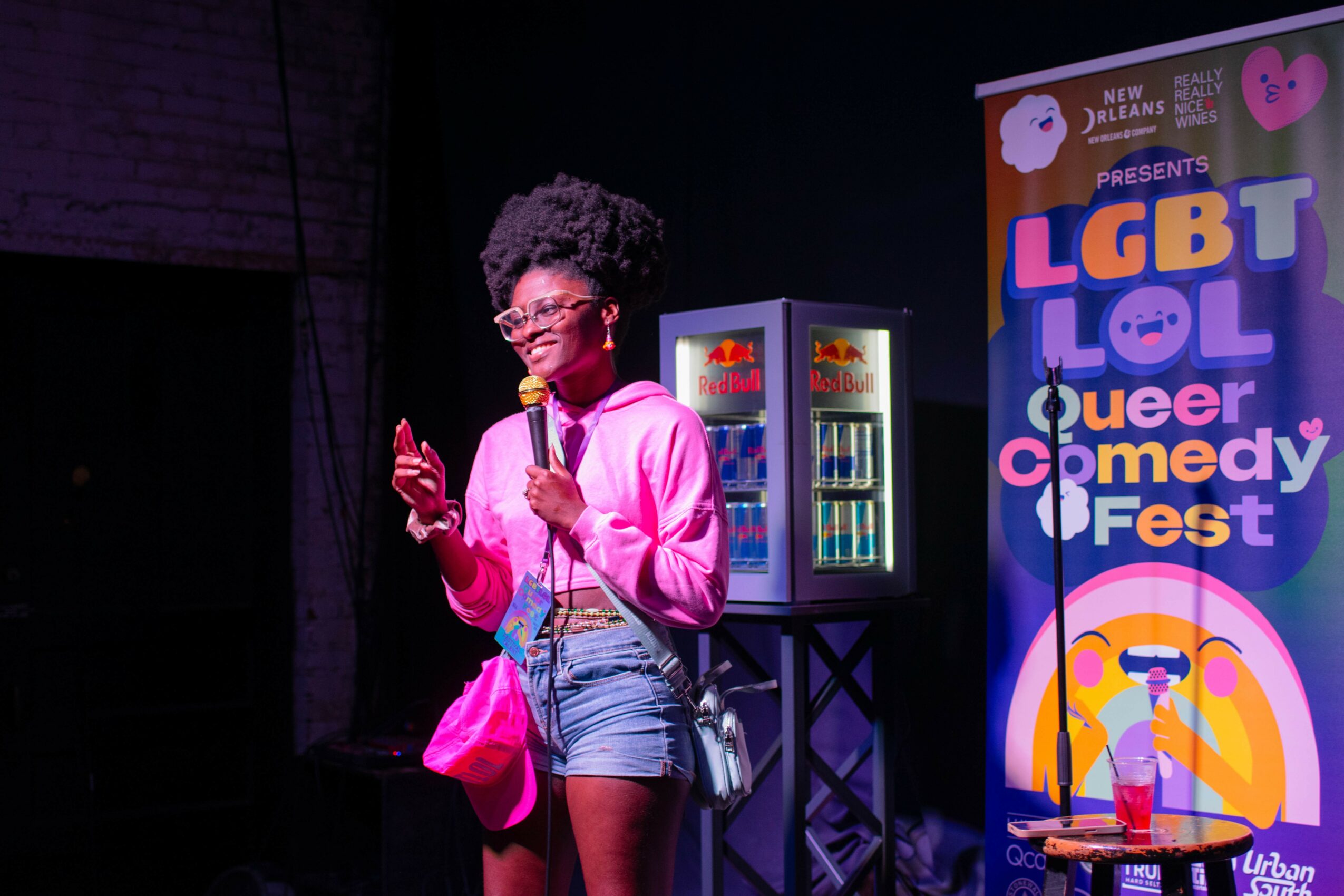 Person with curly hair and glasses speaking into a microphone on stage, colorful LGBT comedy fest banner in background.