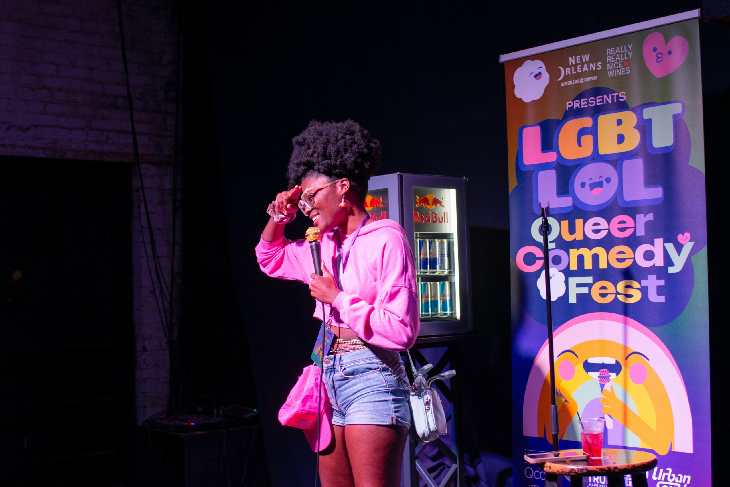 Person with curly hair wearing pink hoodie and denim shorts speaking into a microphone on stage at LGBT comedy event.