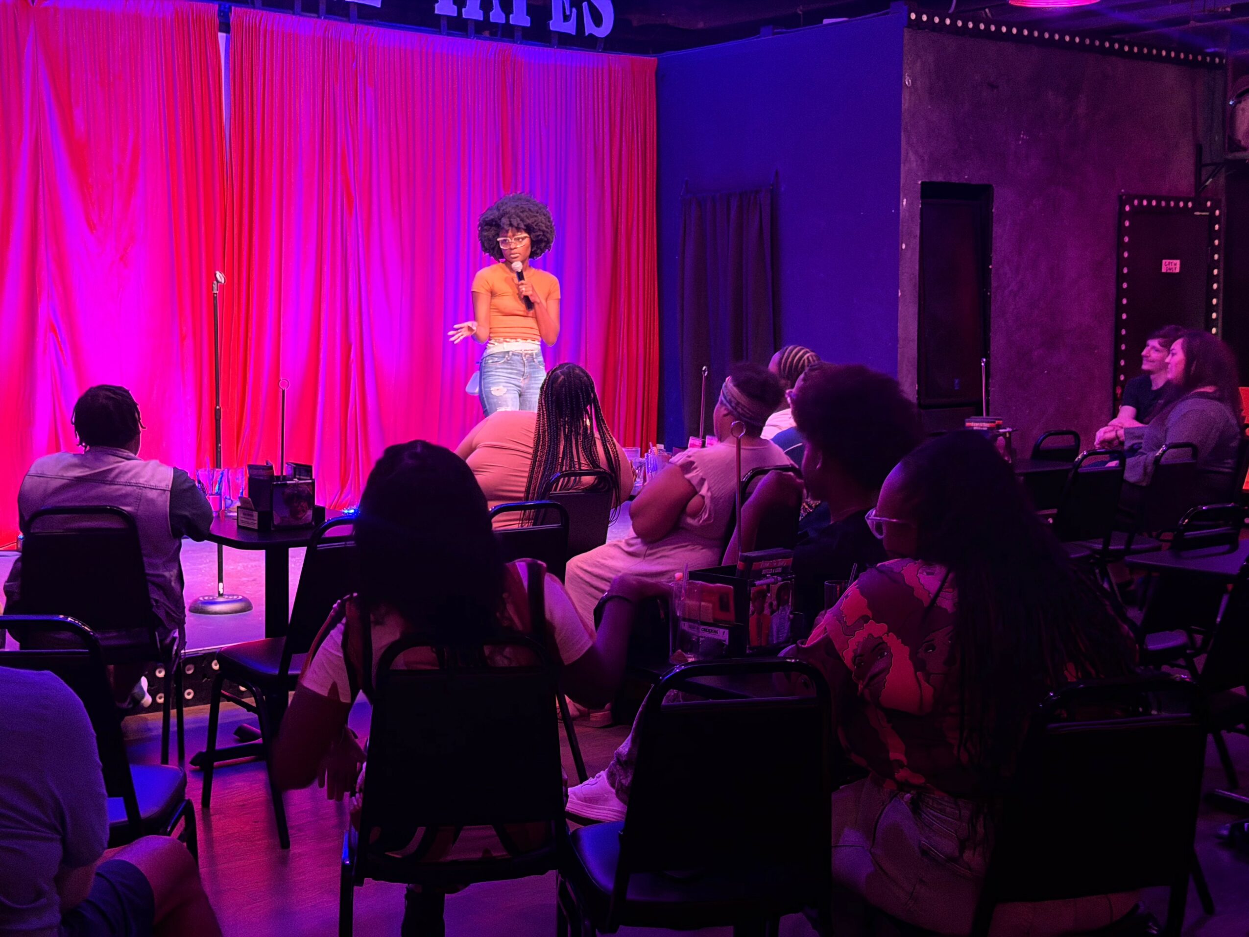 Performer with curly hair on stage in front of pink curtain, audience seated at tables watching, dim lighting.