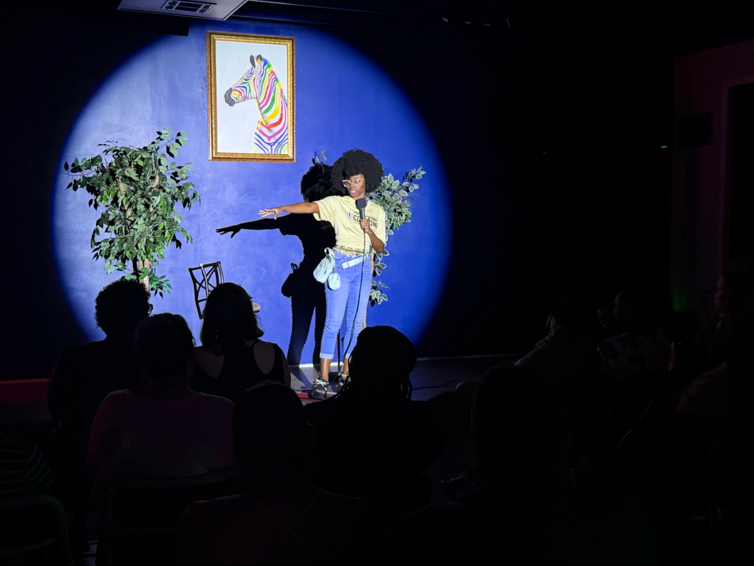 Two performers on stage with a blue background, one with a microphone, surrounded by audience silhouettes.