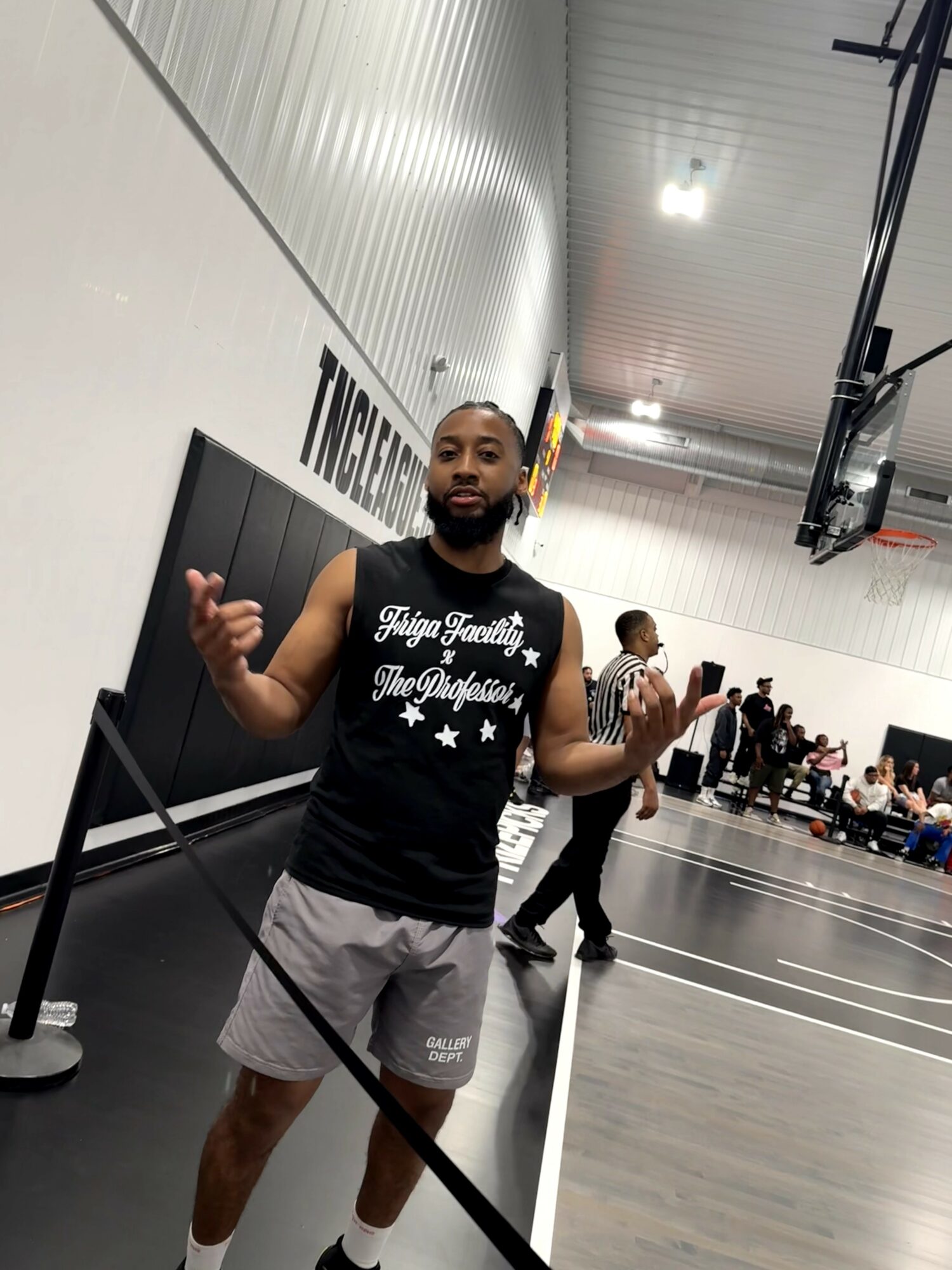 Man with a beard and sleeveless shirt standing on a basketball court, holding a basketball, with people in the background.