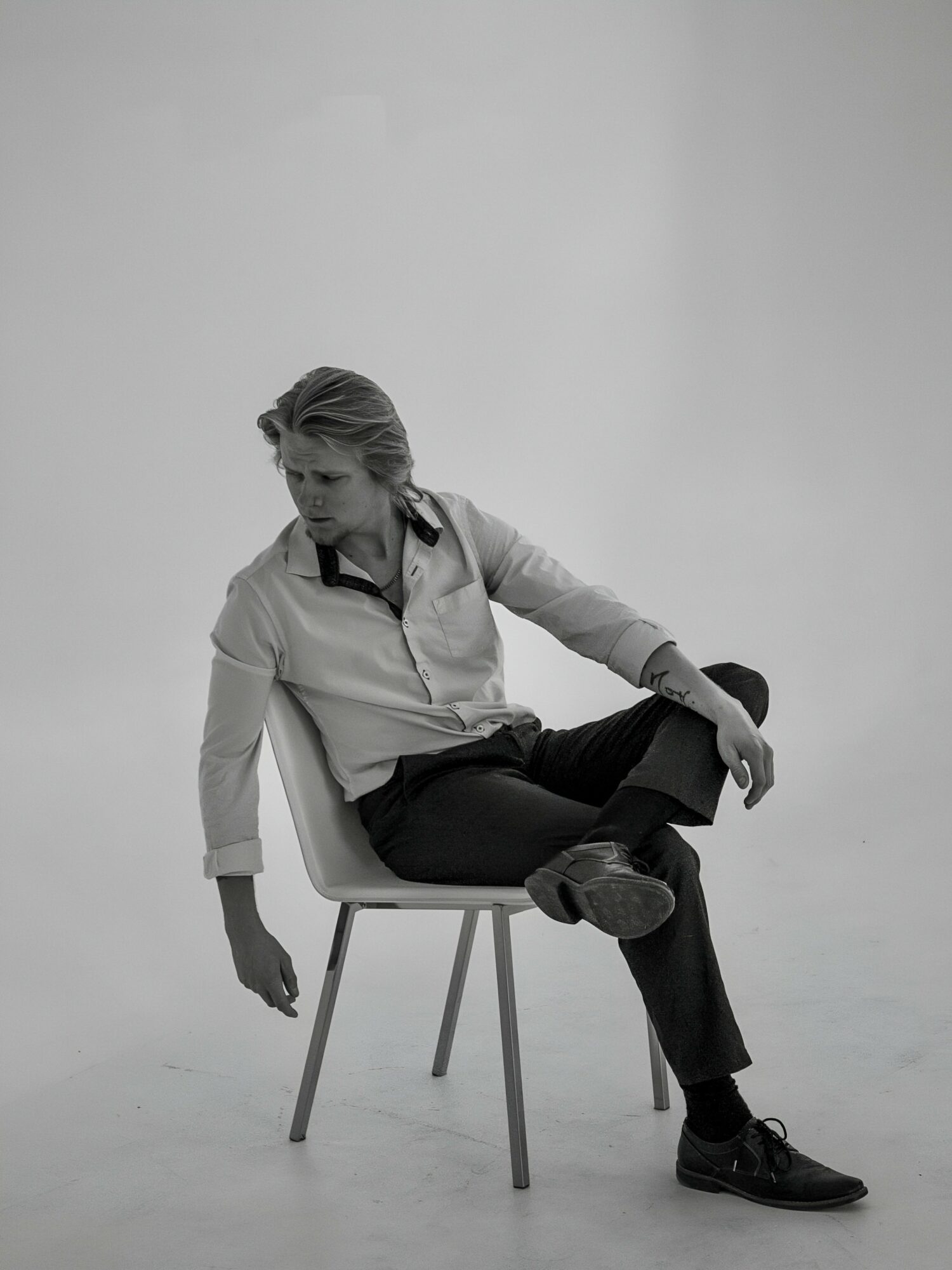 Person sitting sideways on a chair with legs crossed, in a studio setting, looking down, black and white photo.