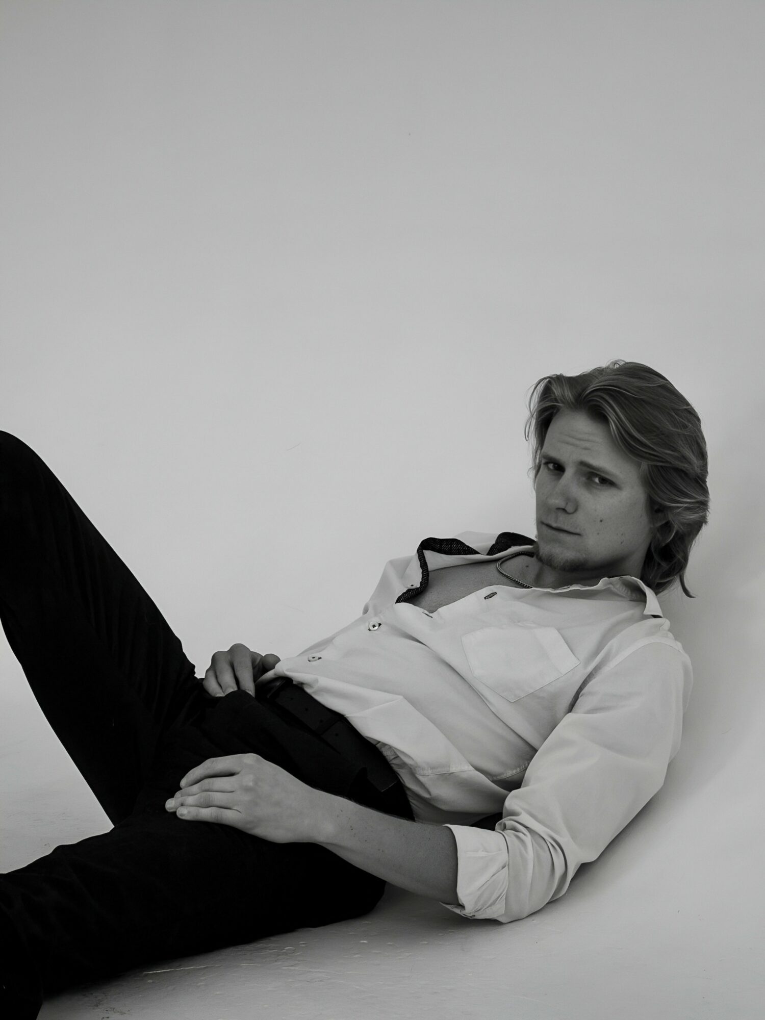 Person with wavy hair lying on the floor, wearing a light-colored shirt and dark pants, looking to the side.
