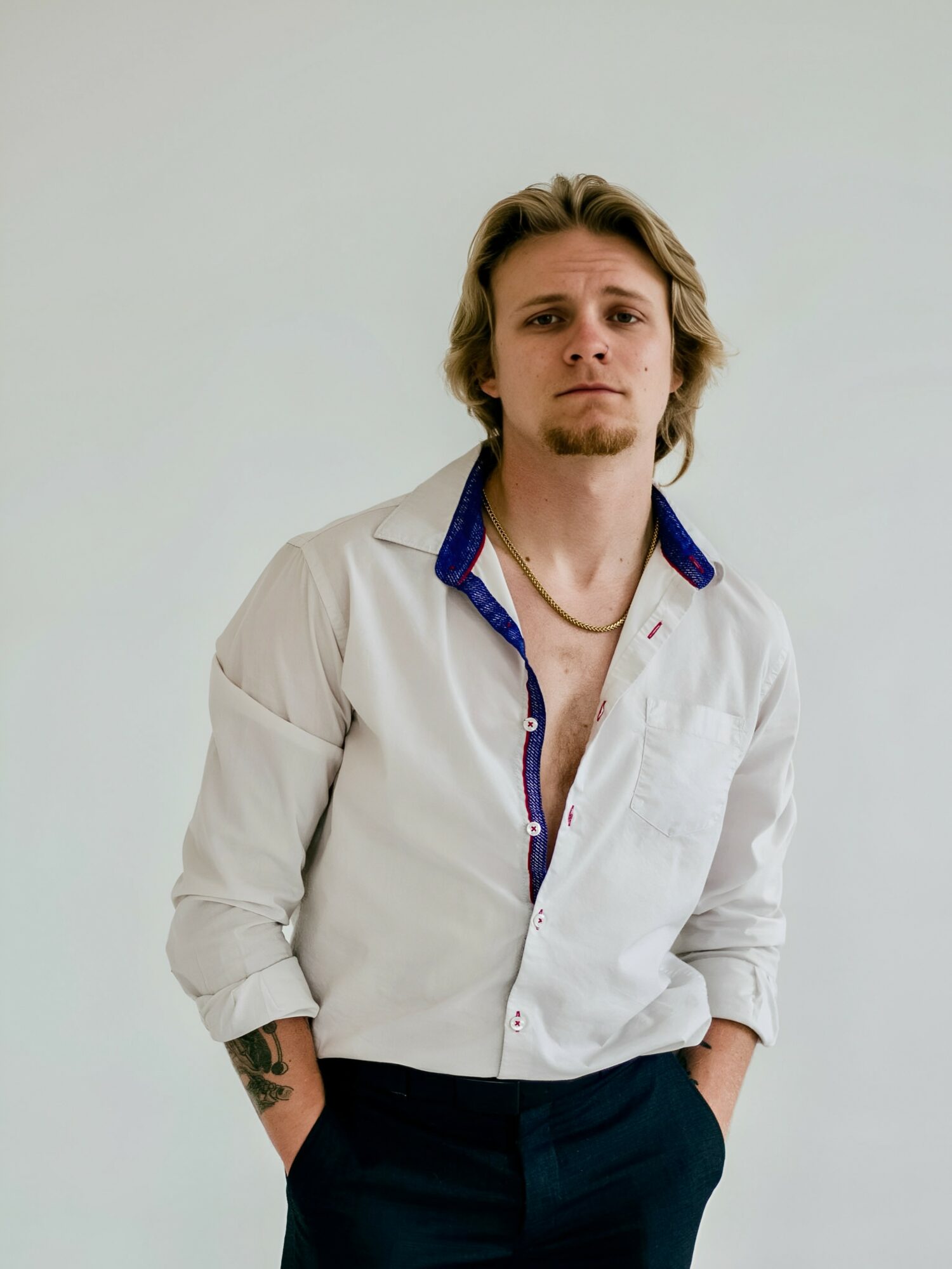 Young man with shoulder-length hair, wearing a partially unbuttoned white shirt with a blue collar, standing with hands in pockets.