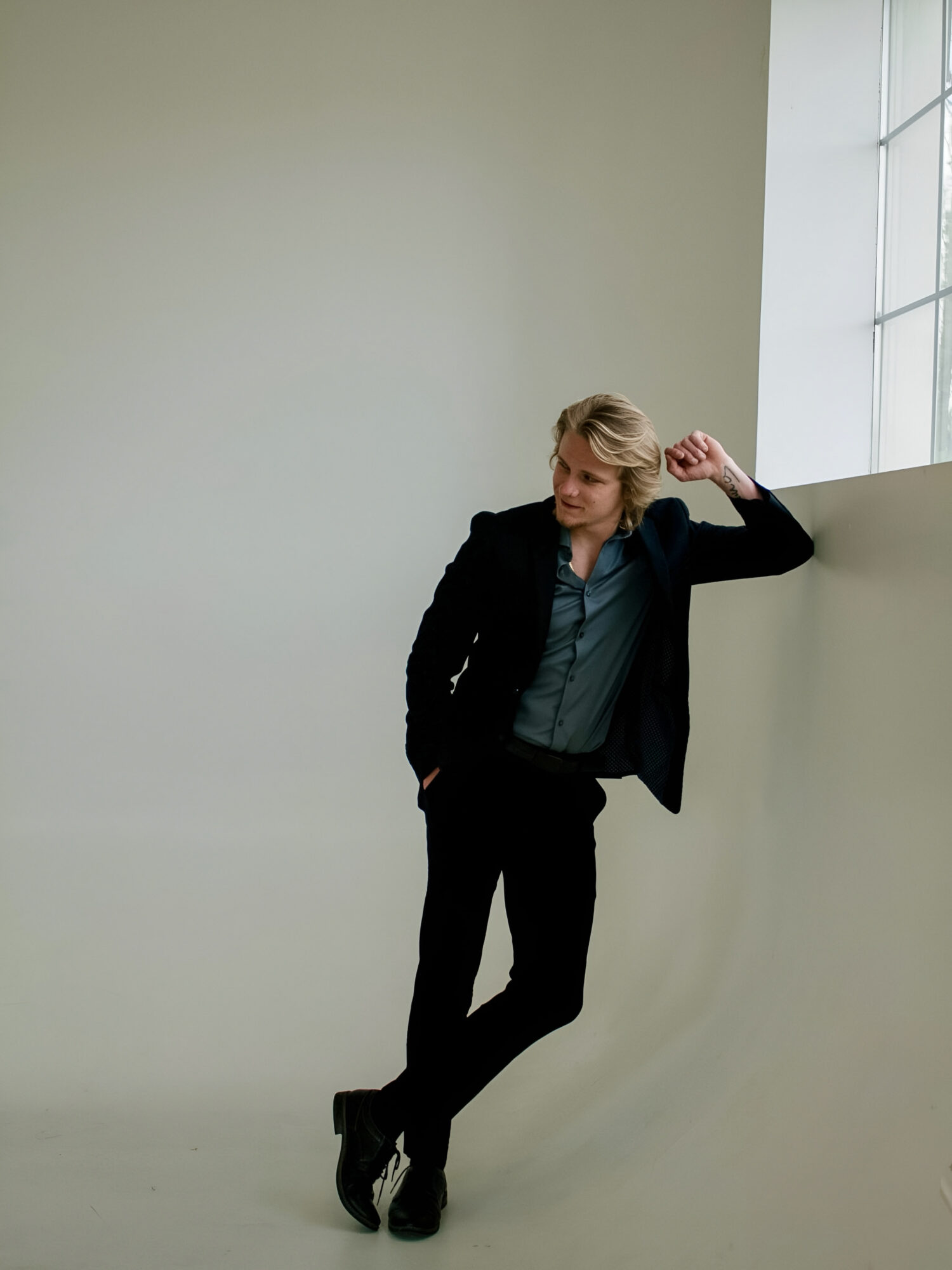 Woman with blonde hair in a black blazer and pants leaning against a wall near a window, looking to the side.