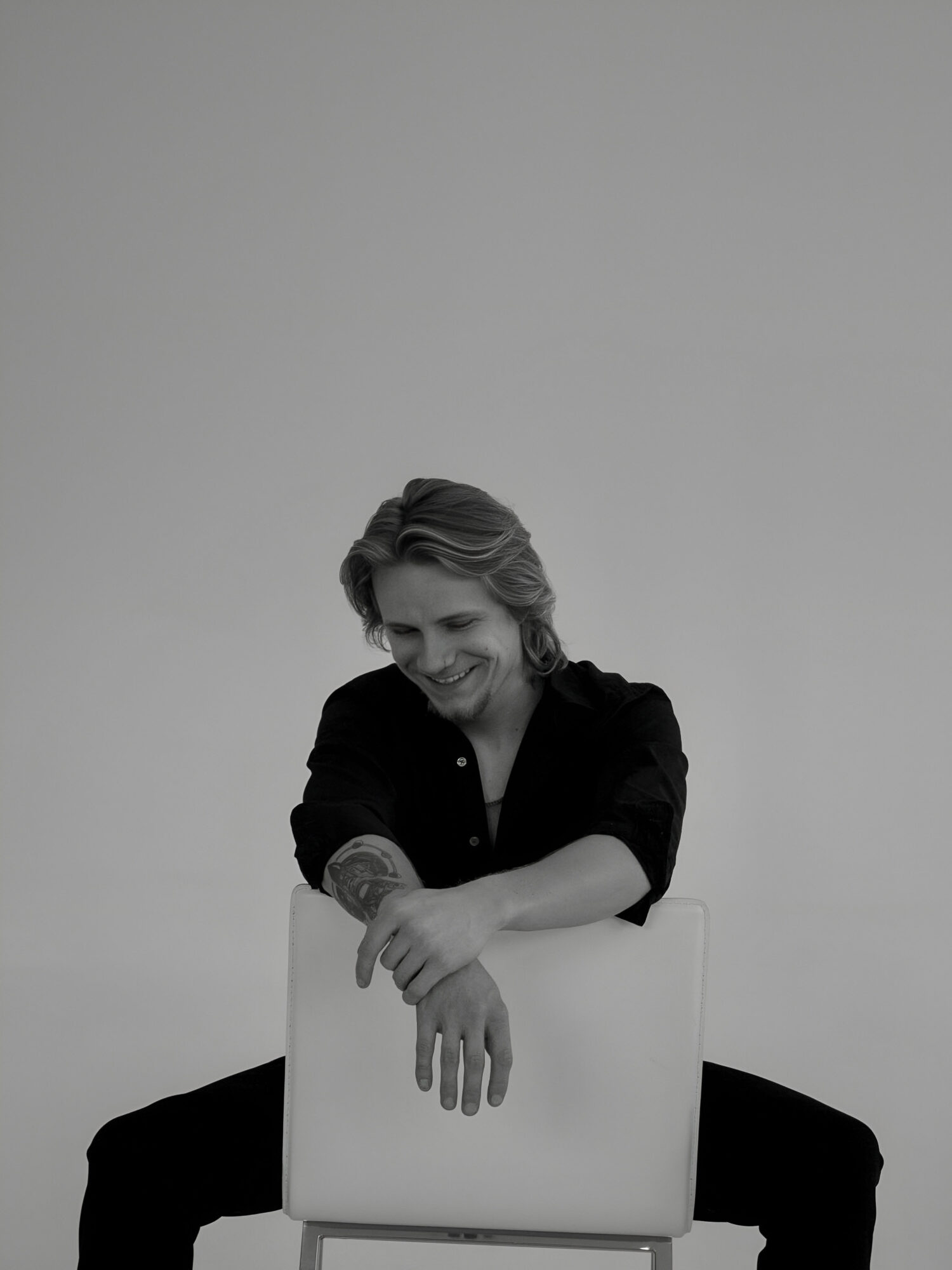 Person with short hair smiling, sitting with legs apart, leaning on a chair, in black clothing, against a plain background.