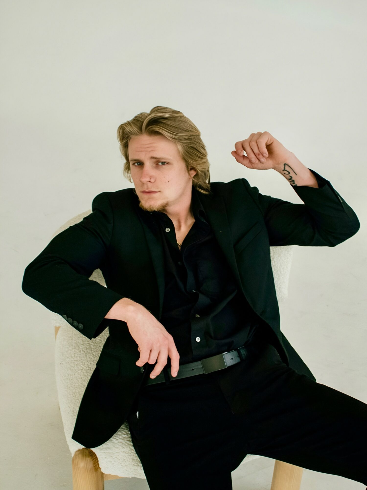 Young man with blonde hair in a black suit, sitting on a chair, raising his right arm, against a plain background.
