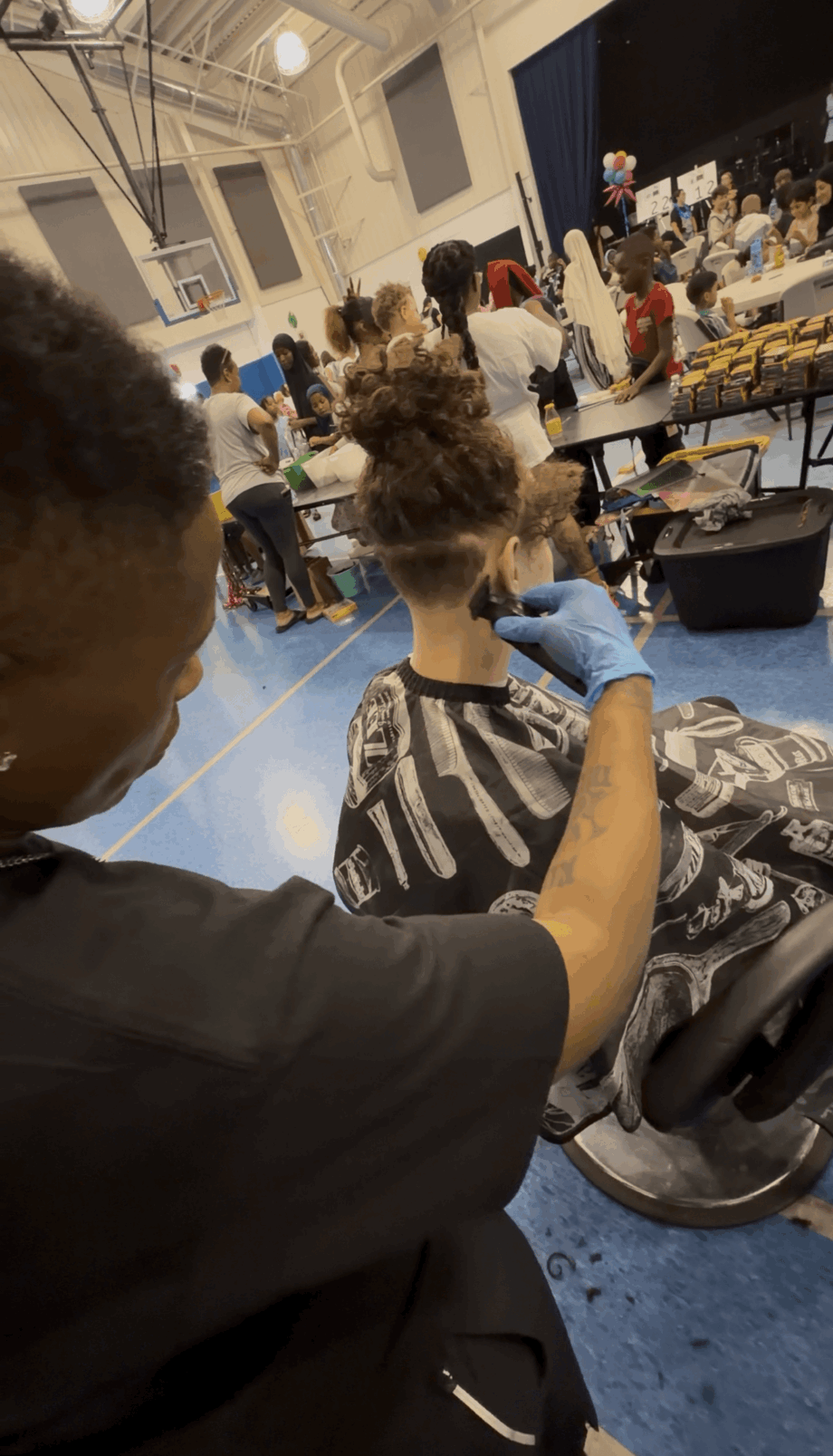 Person with curly hair and gloves working on a mannequin head in a large indoor space with people and tables.