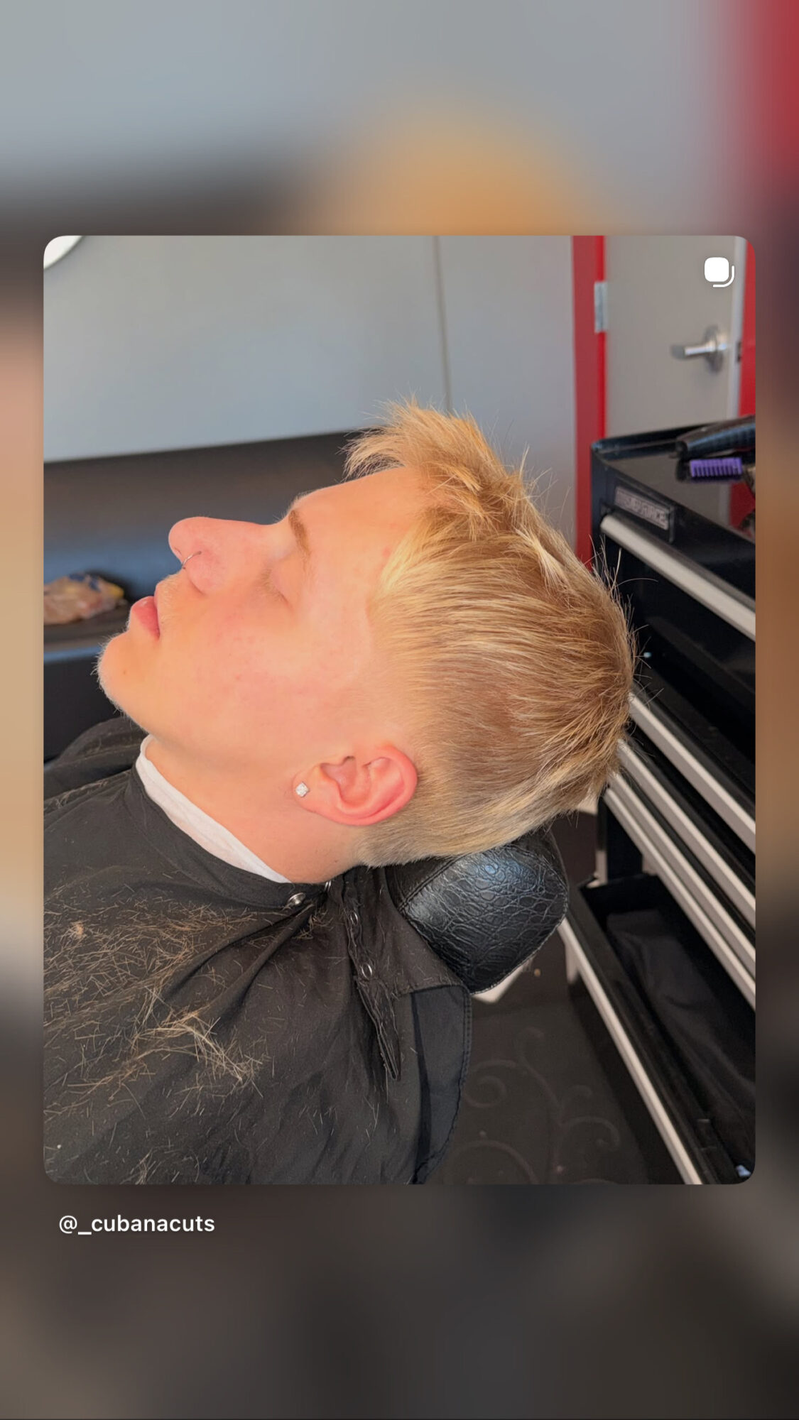 Person with blonde hair lying back in a chair, eyes closed, in a salon or barbershop setting.