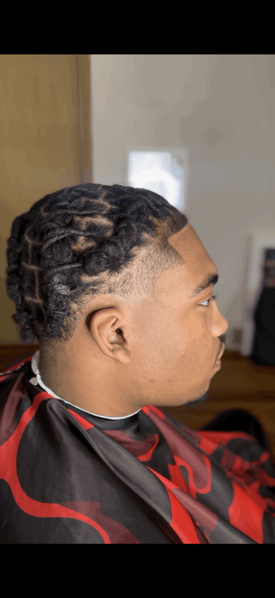 Side view of a person with styled dreadlocks, sitting in a salon chair with a cape, in a room with a mirror and furniture.