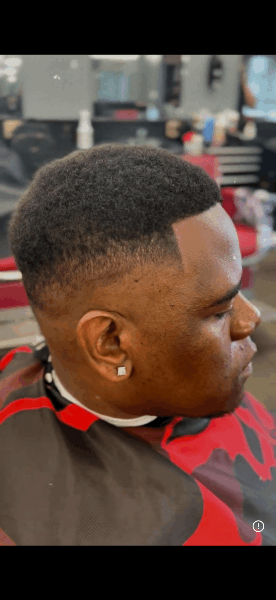 Side profile of a young man with a short, textured haircut, wearing a red and black shirt, in a barbershop.