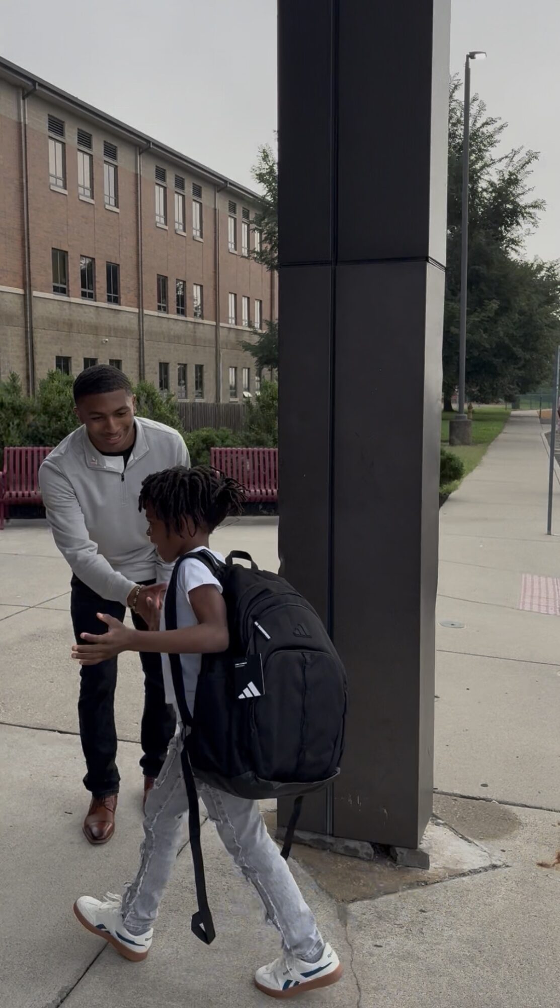 Two children standing on sidewalk near a large black pole, one with a backpack, talking outdoors.