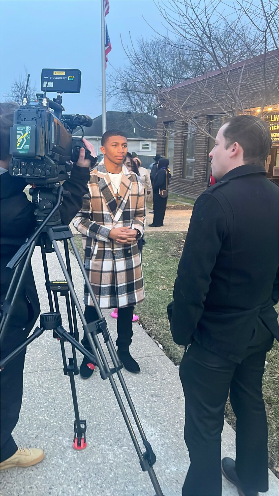 Two people being interviewed outdoors with a camera and crew nearby, trees and a building in the background.